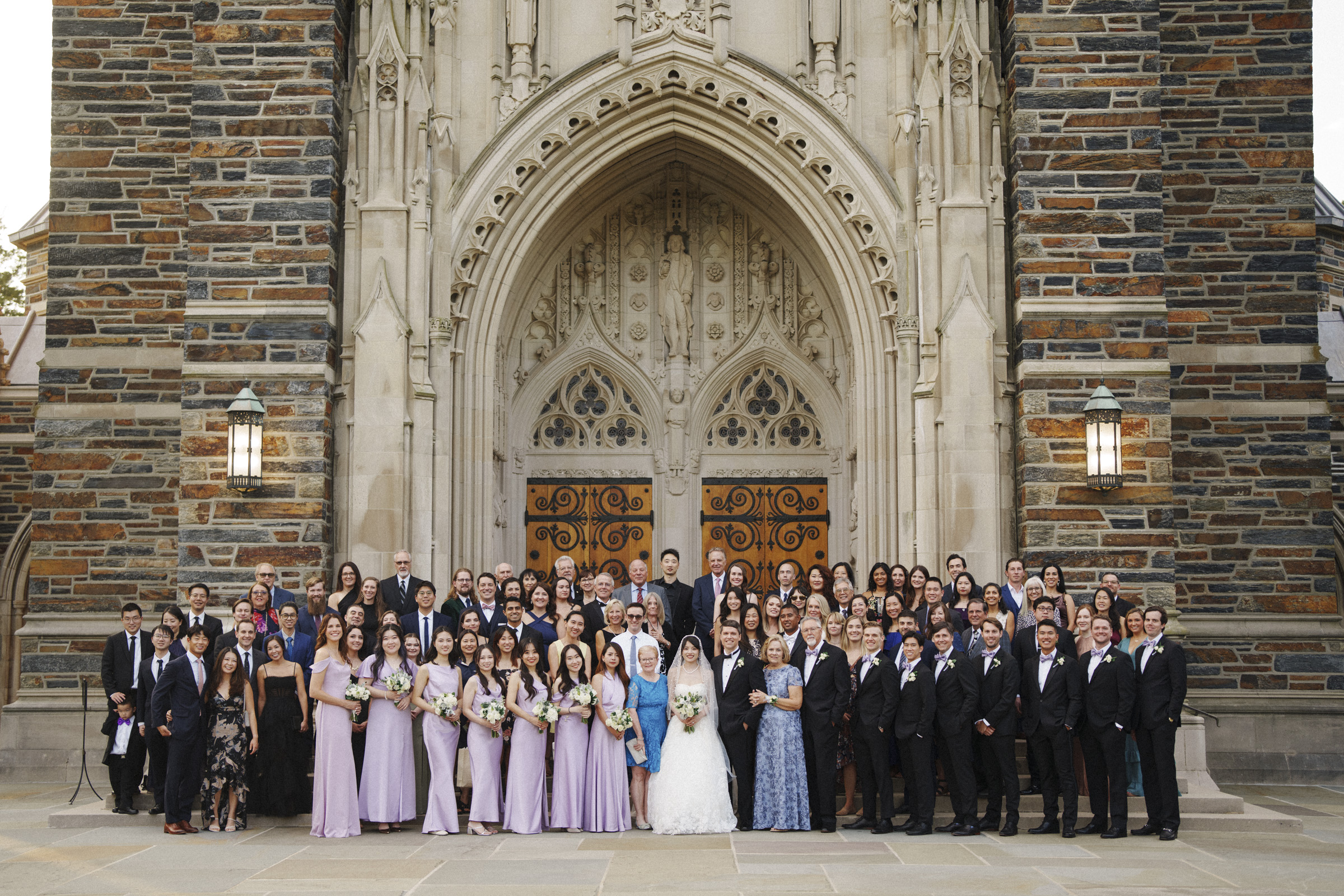 A large wedding group poses in front of a Gothic-style church entrance. The bride and groom stand at the center, surrounded by bridesmaids in lavender dresses and groomsmen in suits, along with other guests. The church features ornate stonework and large wooden doors.