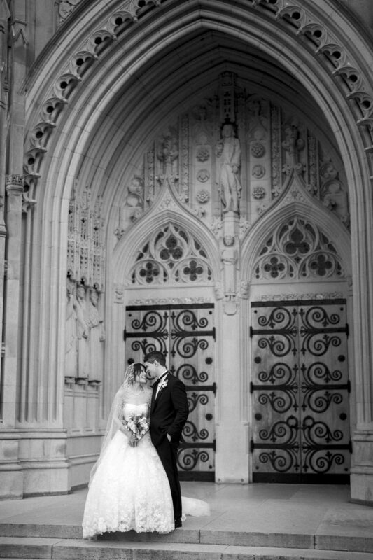 A bride and groom stand in front of an ornate, gothic-style church door. The bride is wearing a long lace gown and veil, holding a bouquet, while the groom is in a suit. They are looking at each other under the archway.