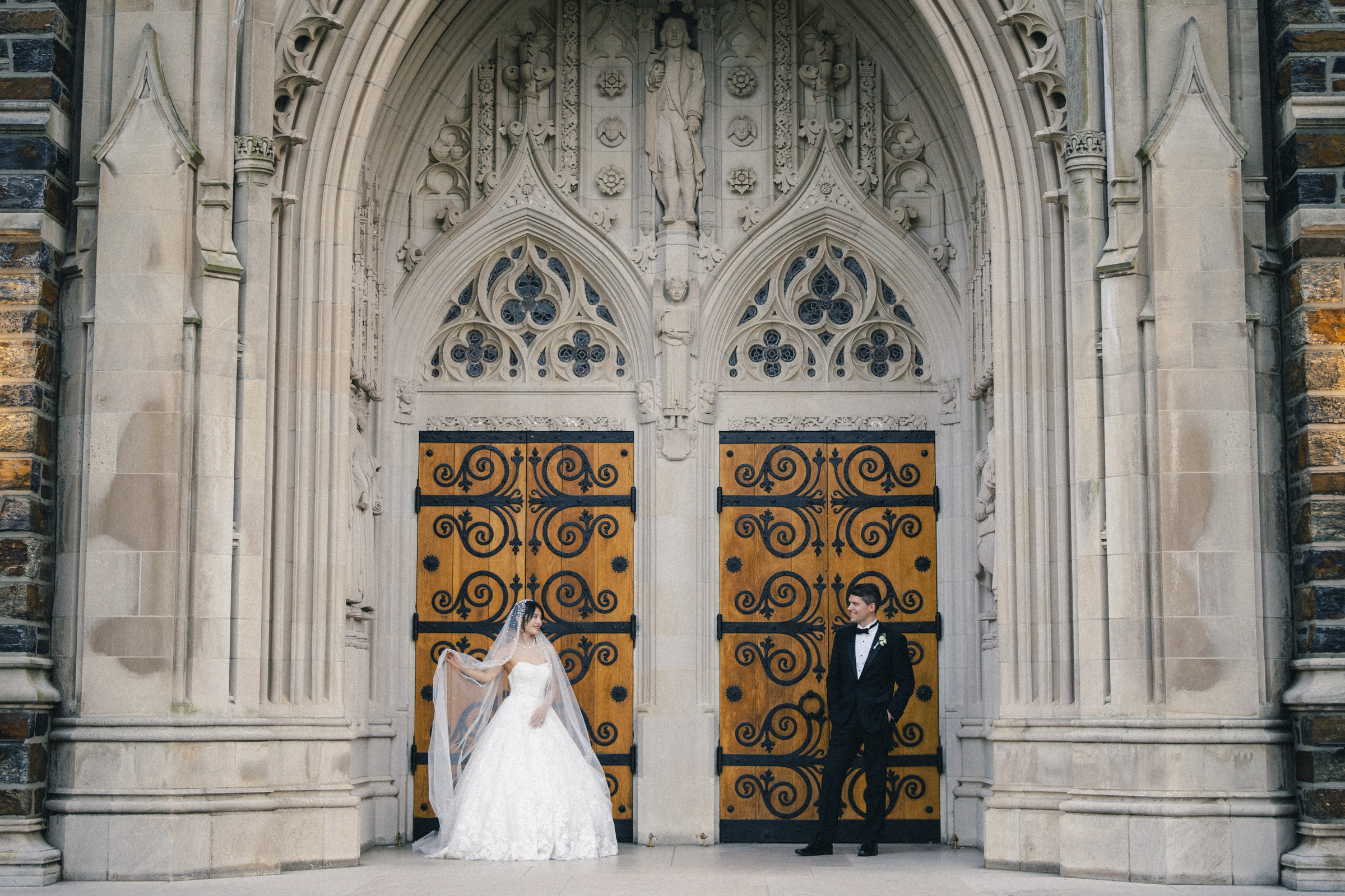 A bride and groom stand in front of an ornate church entrance. The bride, in a white gown, holds a veil as the groom in a black tuxedo looks on. The wooden doors are intricately carved, framed by grand stone archways.