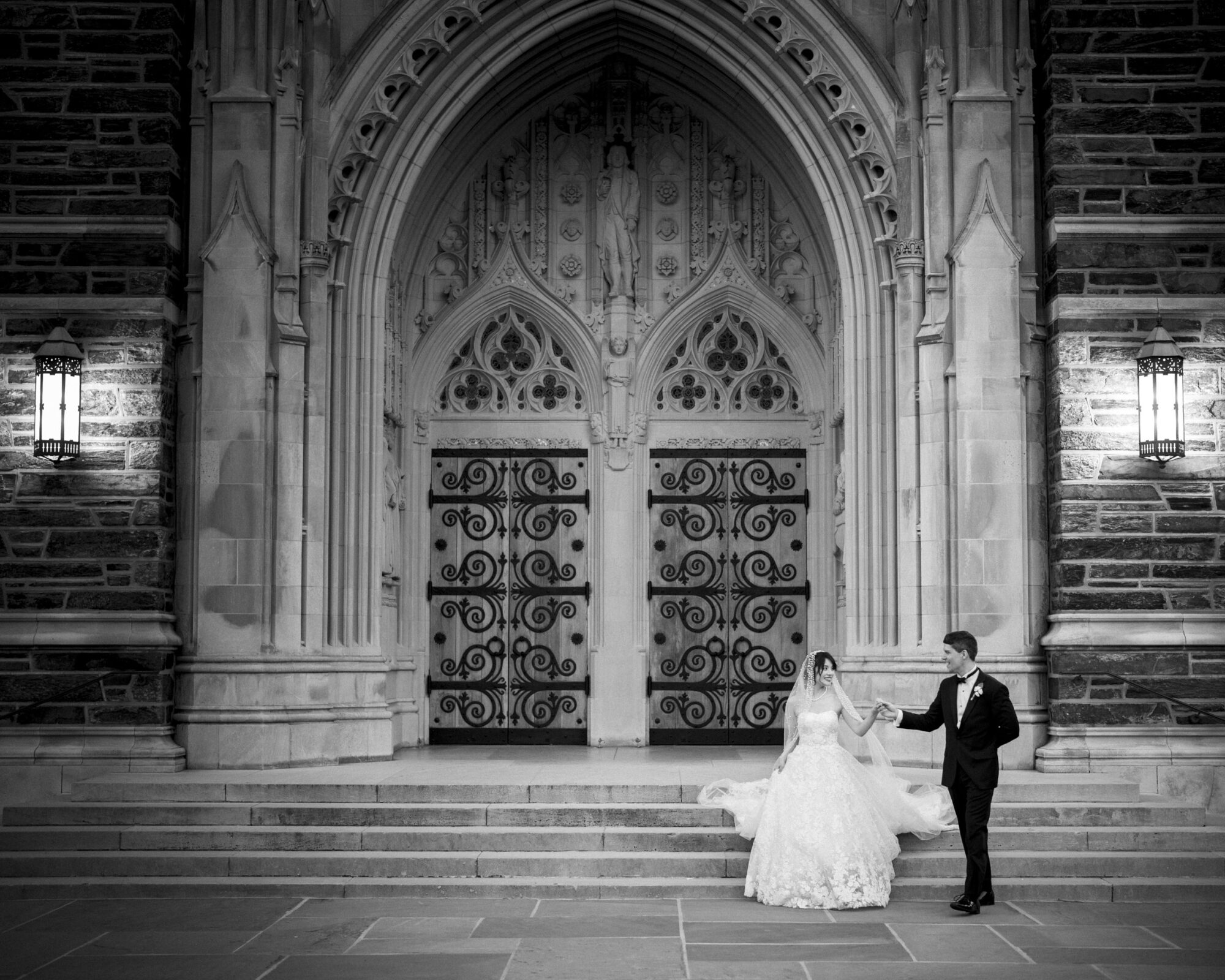 A bride and groom pose on the steps of a grand, ornate church entrance. The groom, in a suit, holds the brides hand as she wears a flowing gown. Intricate stonework and decorative doors are illuminated by lanterns. Black and white photo.