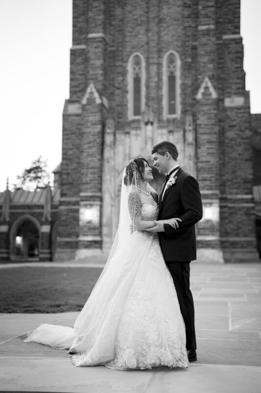 A bride and groom gaze lovingly at each other in front of a tall, intricately detailed stone building. The bride wears a lace gown and veil, while the groom is in a suit. The black-and-white image captures a romantic, elegant moment.