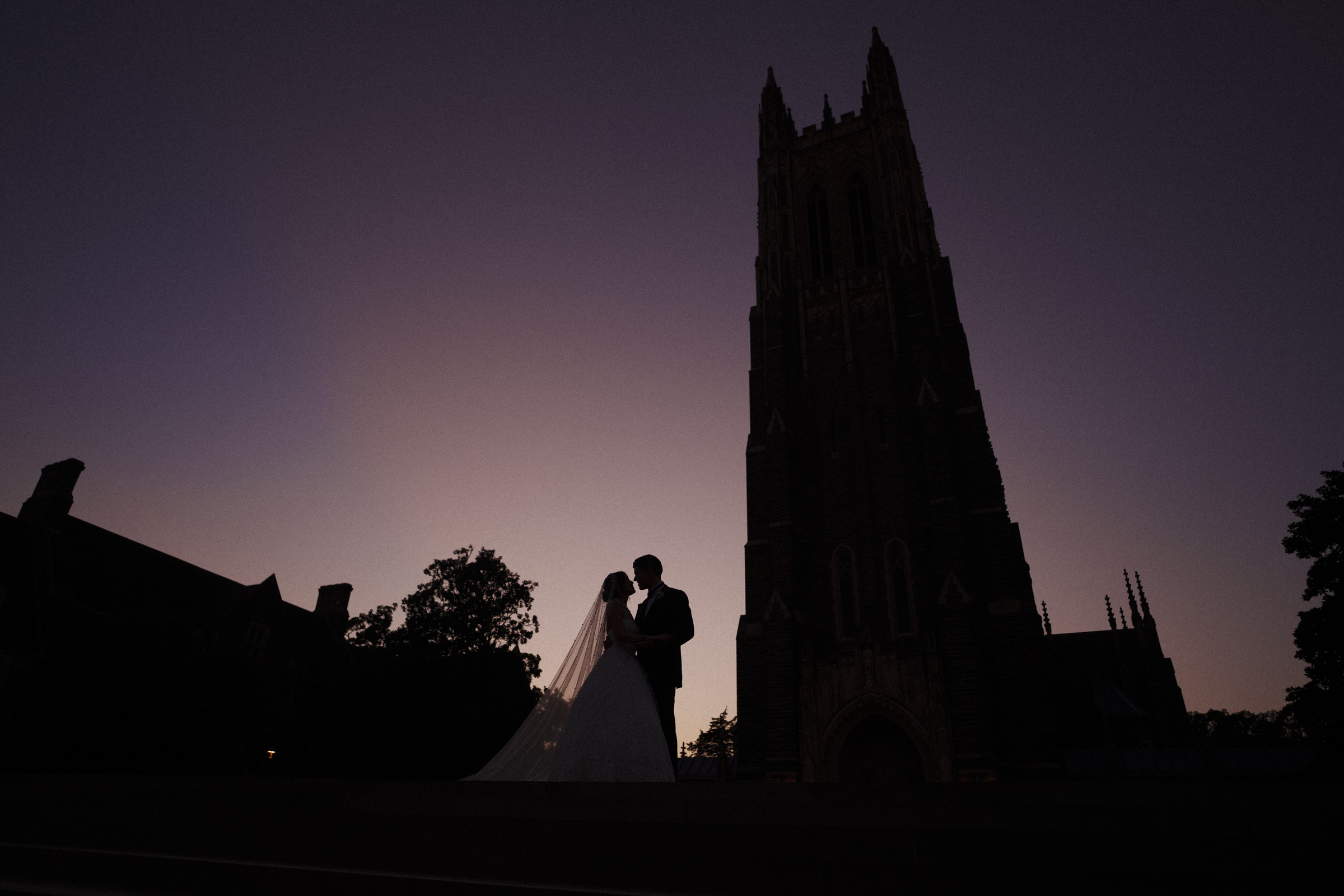 Silhouetted couple in wedding attire embracing at dusk, standing near a tall, gothic-style tower. The sky is softly lit with shades of purple and orange, creating a romantic and serene atmosphere.