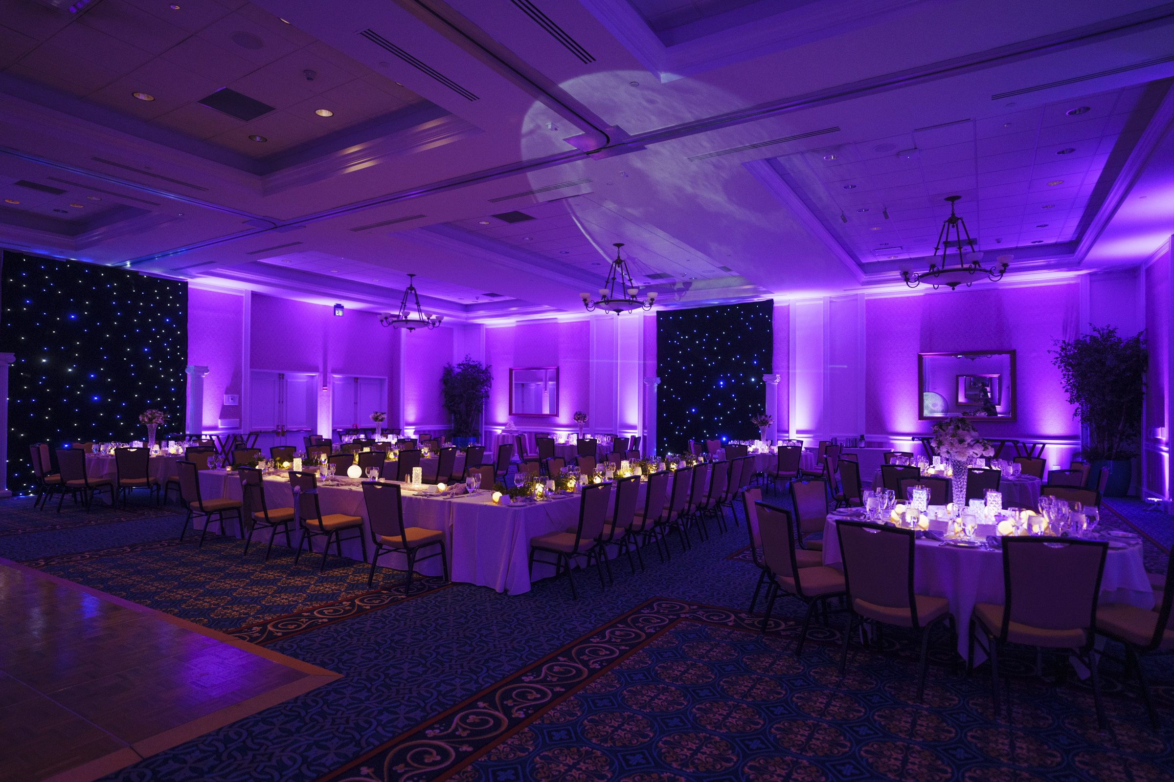 A banquet hall with purple ambient lighting features long and round tables elegantly set for dining. Candles and floral centerpieces adorn the tables. The ceiling is illuminated with a purple moon projection, creating a sophisticated atmosphere.