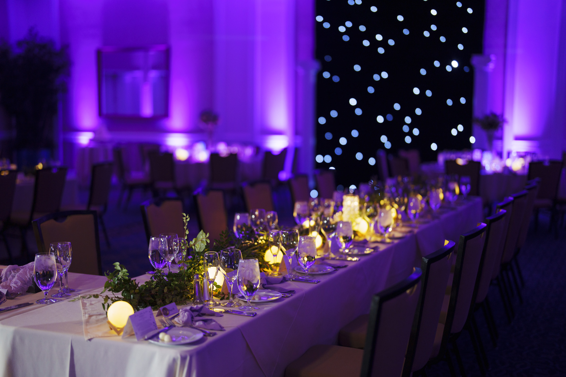 A dimly lit banquet hall with long, elegantly set tables. Table decorations include glassware, foliage, and glowing spherical lights. Purple ambient lighting casts a warm glow, and twinkling white lights adorn a backdrop at the rooms rear.