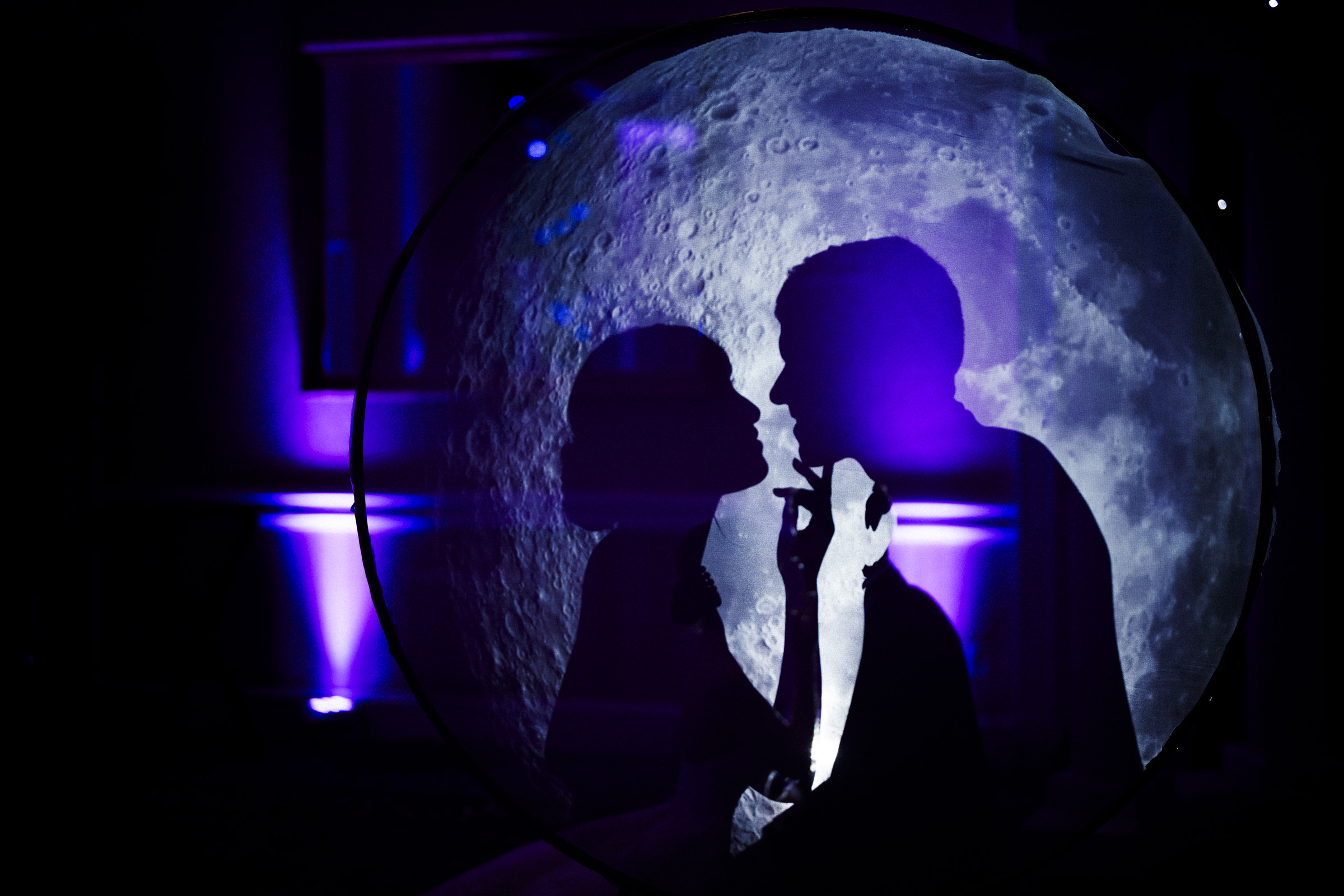 Silhouettes of a couple in front of a large moon backdrop. The scene is illuminated with blue and purple lights, creating a romantic and dreamy atmosphere.