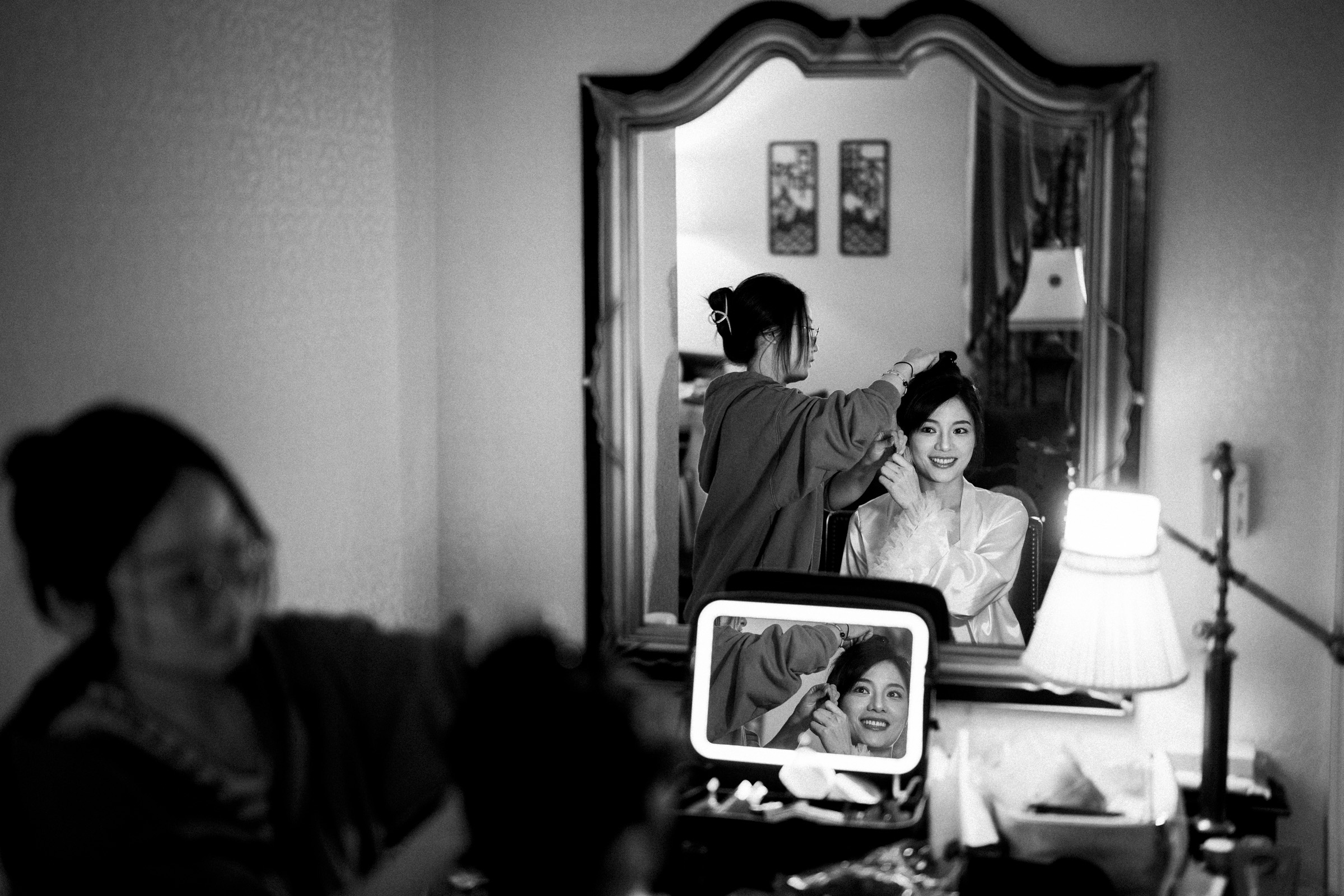 A woman sits in front of a mirror while getting her hair styled. She is smiling at her reflection. The room is softly lit, and there are lamps and small decor items around the mirror. The scene is captured in black and white.