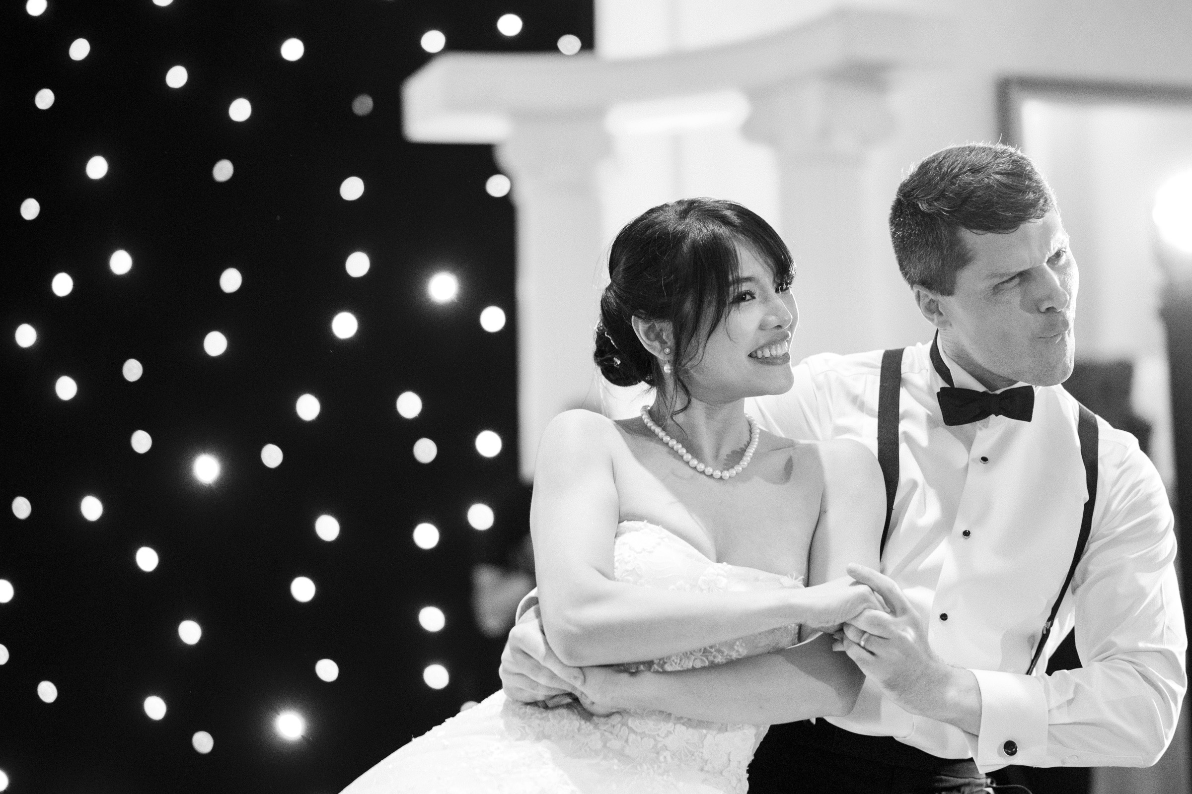 A bride and groom joyfully dance together. The bride is in a strapless gown and pearl necklace, and the groom is in a bow tie and suspenders. They are surrounded by a backdrop of twinkling lights, creating a romantic atmosphere.