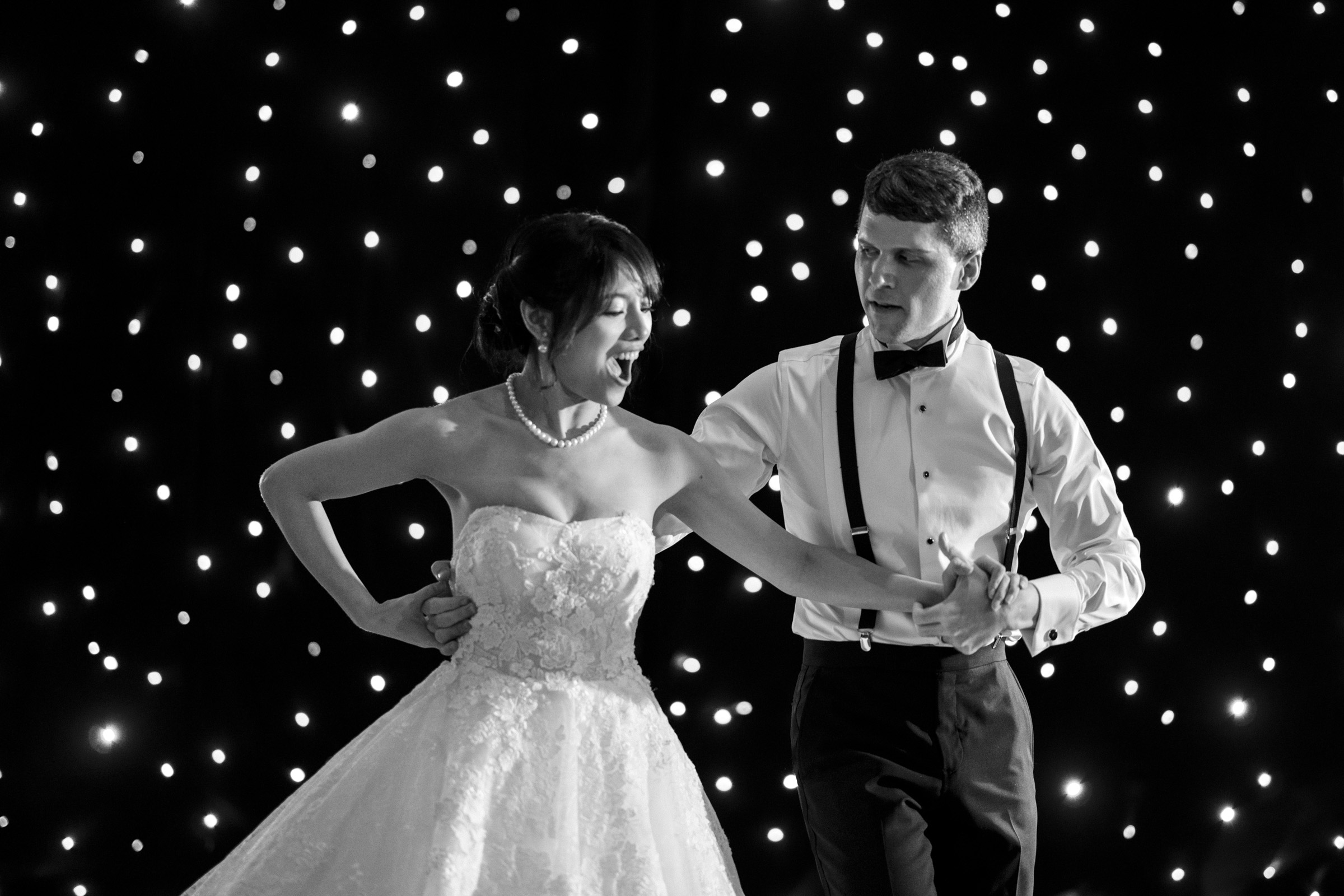 A bride and groom dance joyfully against a backdrop of sparkling lights. The bride wears a strapless gown, and the groom is in a white shirt with suspenders. Both appear happy and engaged in their dance.