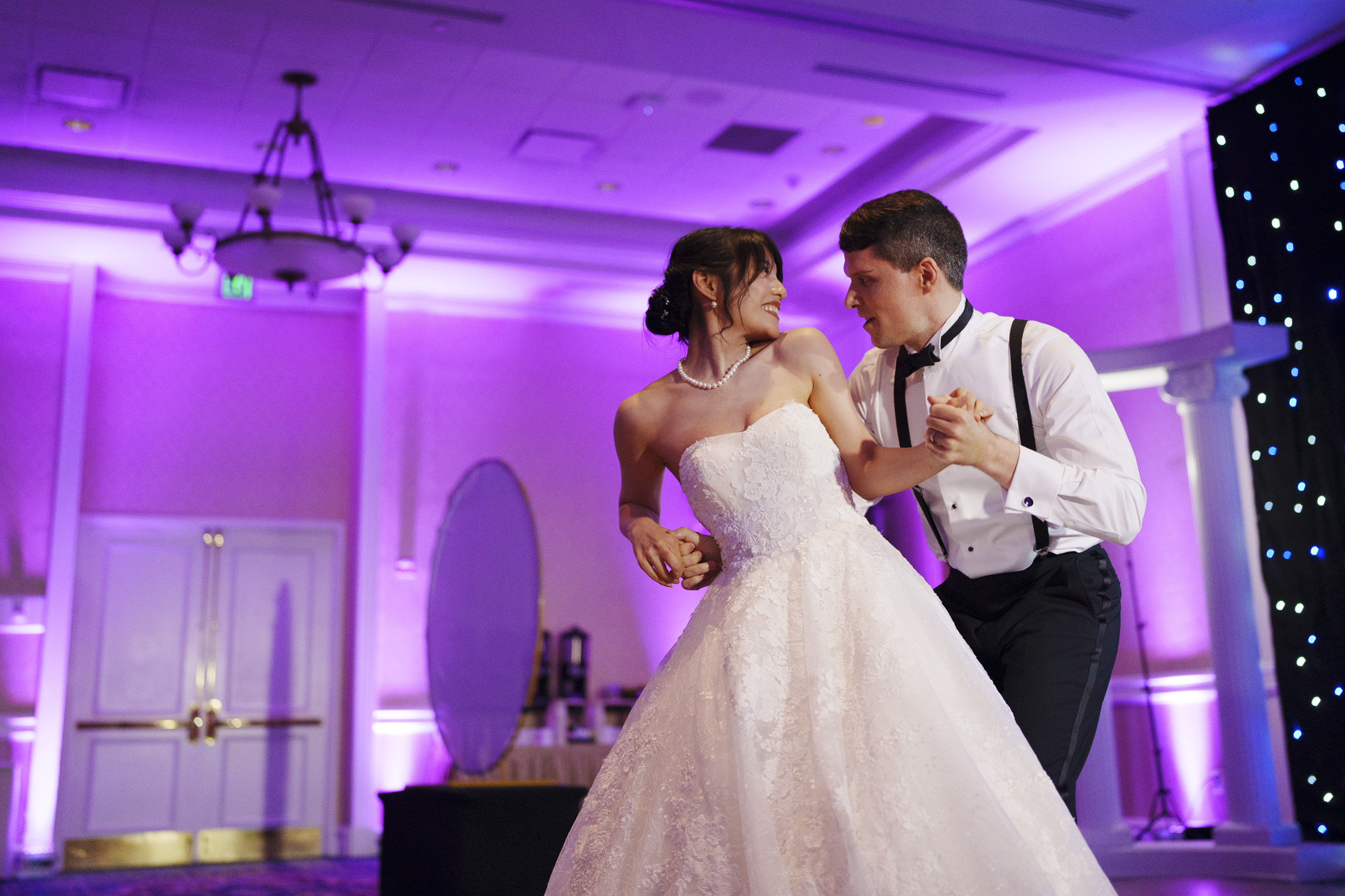 A bride and groom hold hands and smile at each other while dancing in a beautifully lit room with purple ambient lighting. The bride wears a white gown, and the groom is in a white shirt and suspenders.