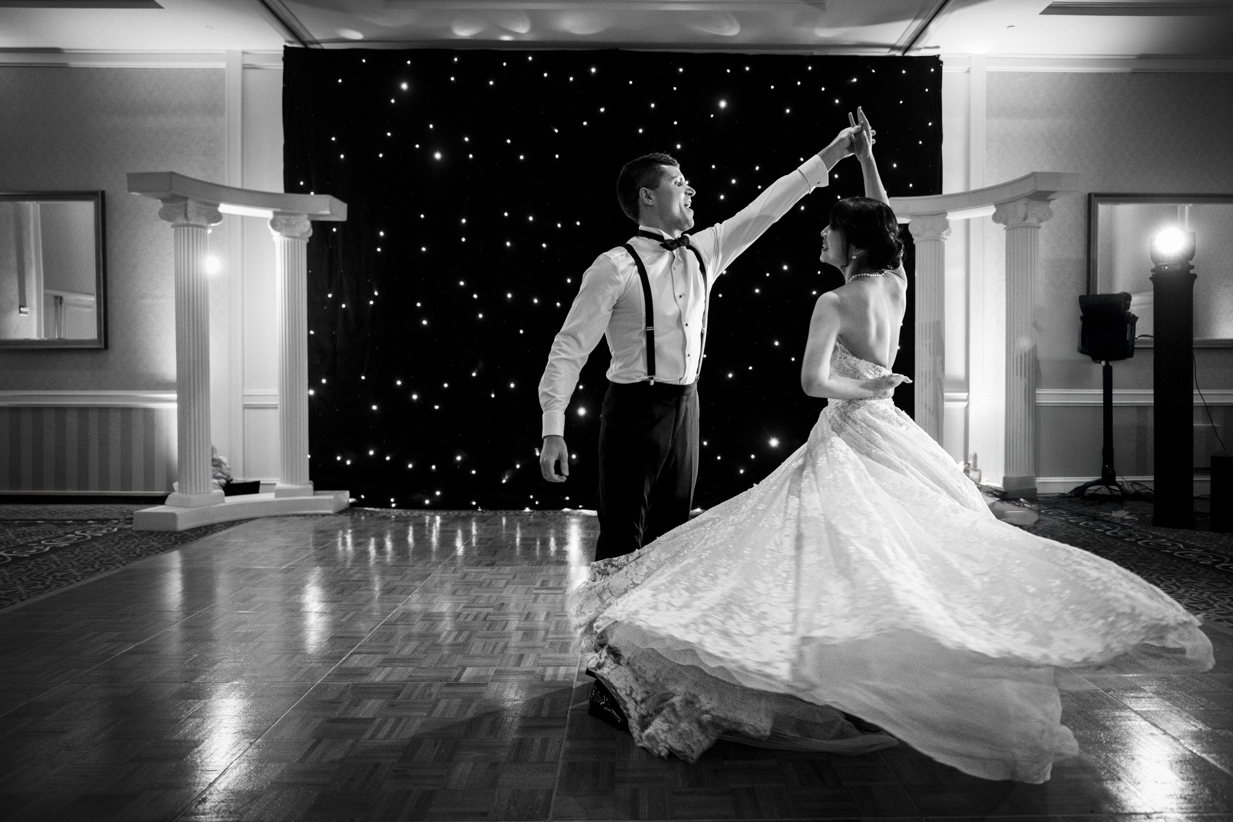 A couple elegantly dances on a polished wooden floor in a ballroom. The man wears a shirt and suspenders, and the woman wears a flowing gown. They twirl in front of a starry backdrop, with a photographer capturing the moment.