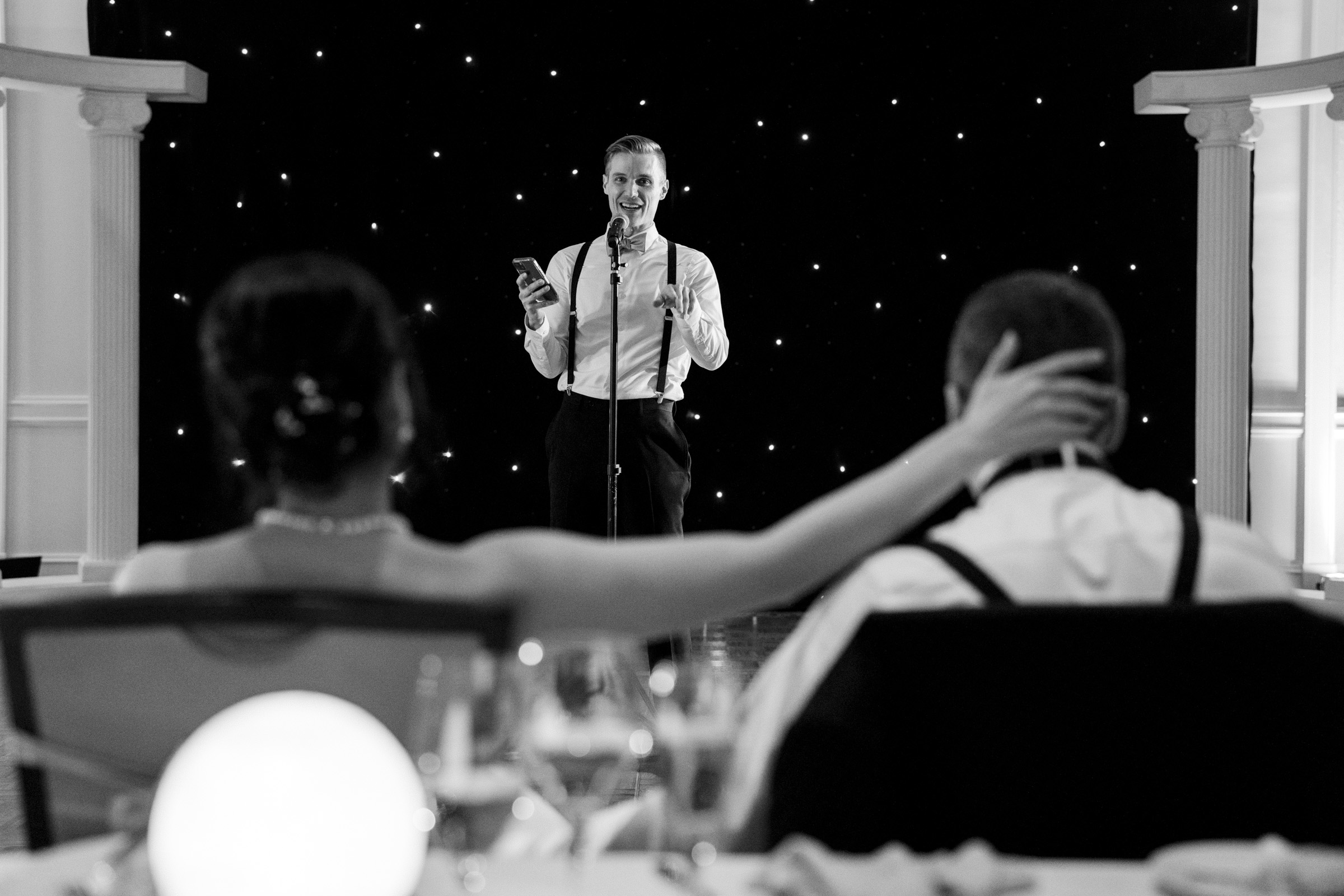 A person giving a speech at a formal event, holding a microphone and a phone. In the foreground, a seated person gently places a hand on the back of another seated person. The background is decorated with glowing star-like lights.