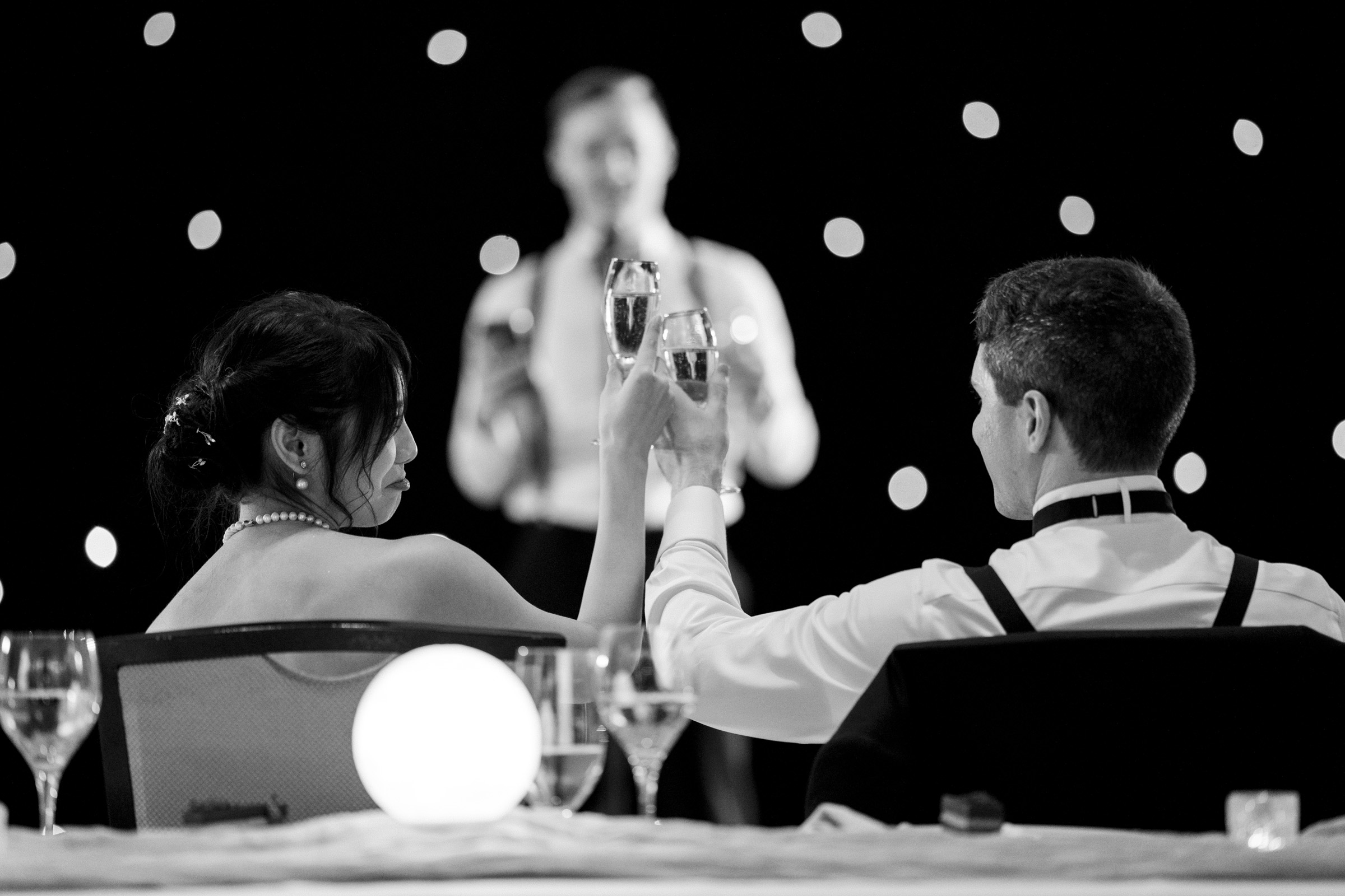 A couple in formal attire clinks champagne glasses while sitting at a table. They are watching a person in the background who appears to be giving a speech. The setting is elegant with dim lighting and small bright lights in the background.