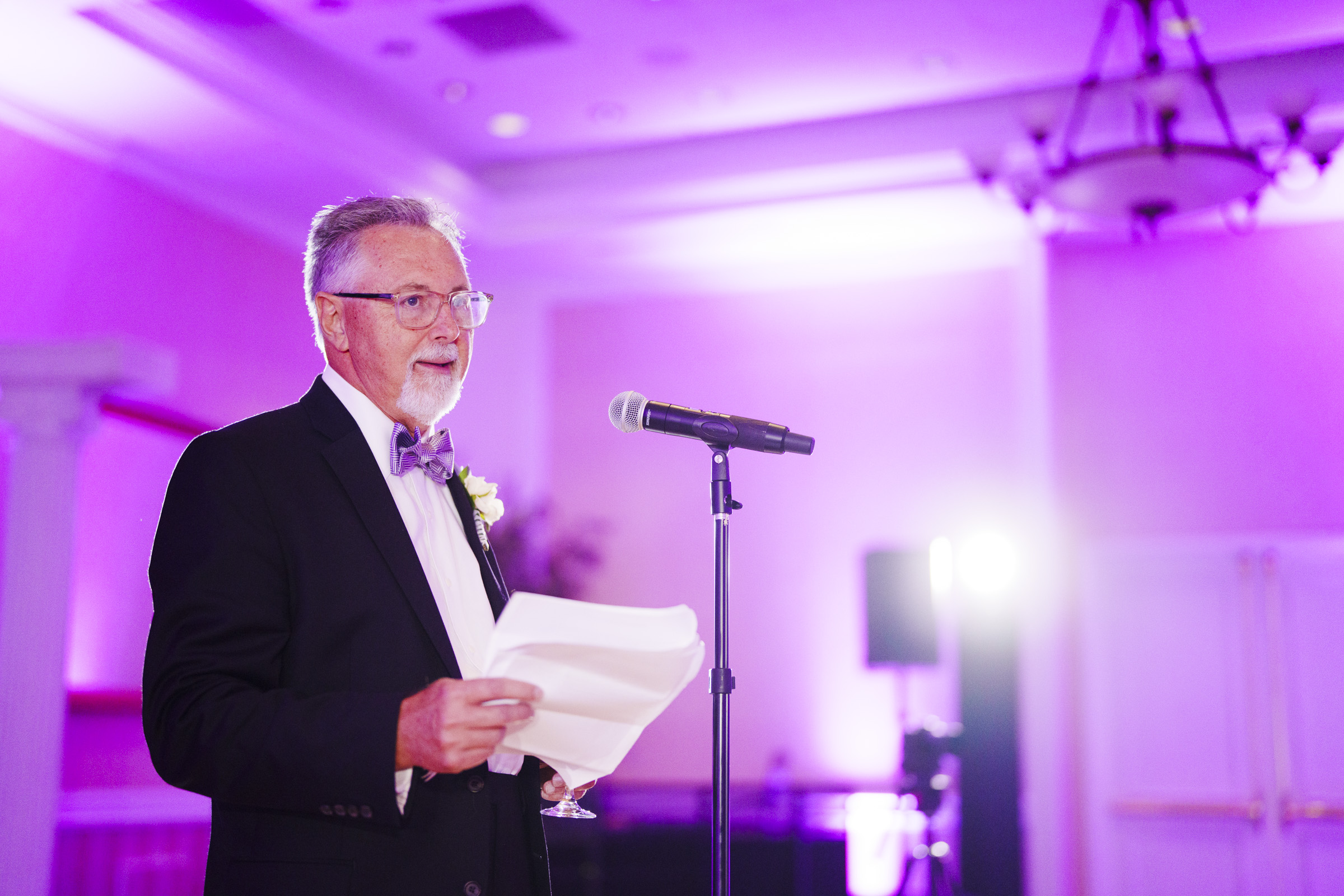 An older man in a suit and bow tie stands at a microphone holding papers. He is speaking in a softly lit room with purple lighting, creating a formal and elegant atmosphere.