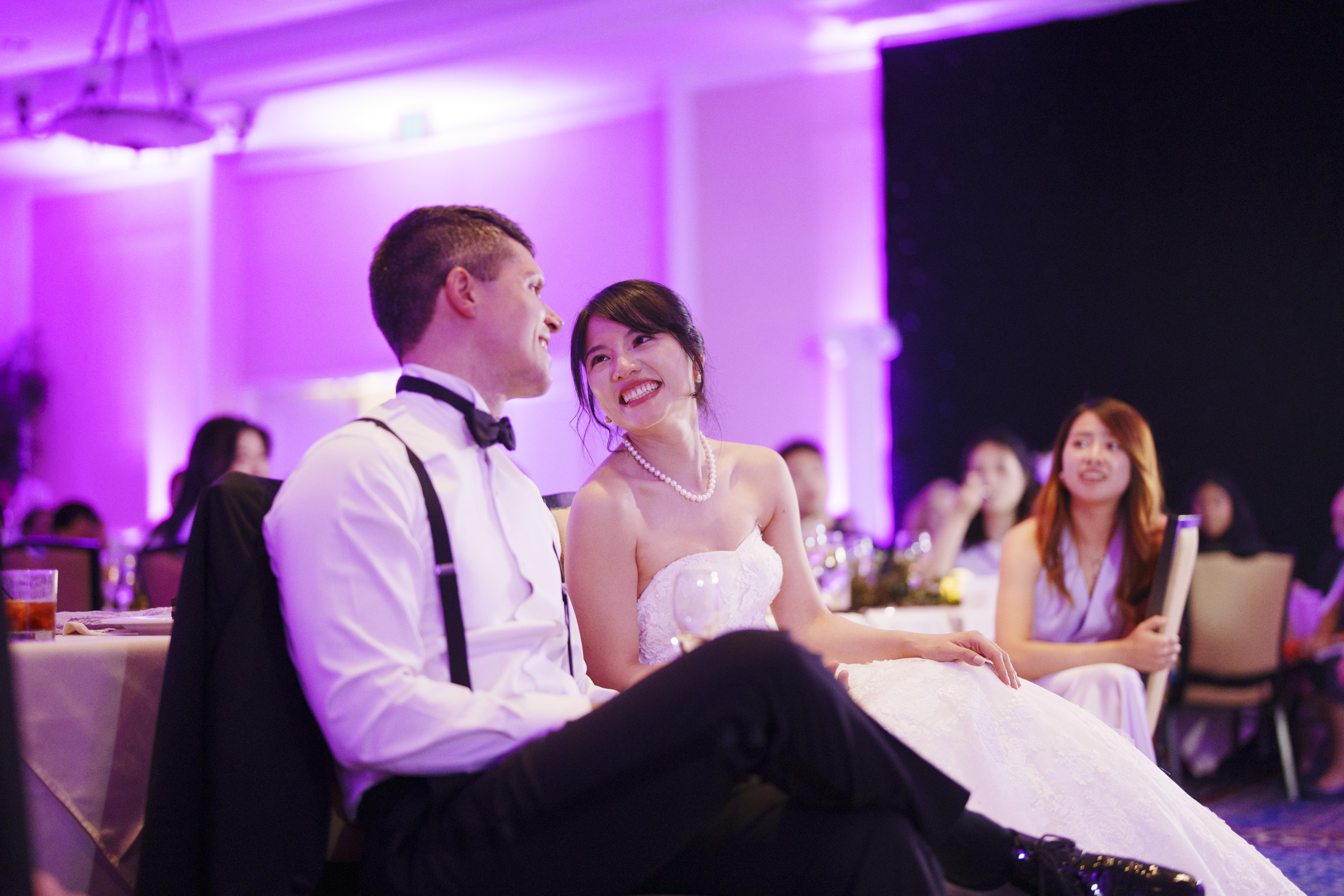 A bride and groom sit closely together, smiling at each other in a warmly lit reception hall. The groom is in a white shirt and black suspenders, while the bride wears a strapless white gown. Guests sit at tables in the softly lit background.