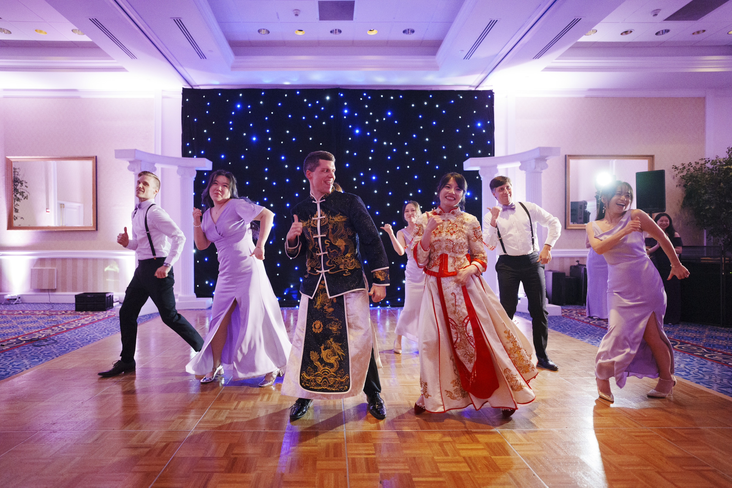 A group of people in traditional and formal attire dance energetically on a wooden floor, against a backdrop of twinkling star-like lights. Everyone appears joyful, with some dressed in matching light-colored outfits.