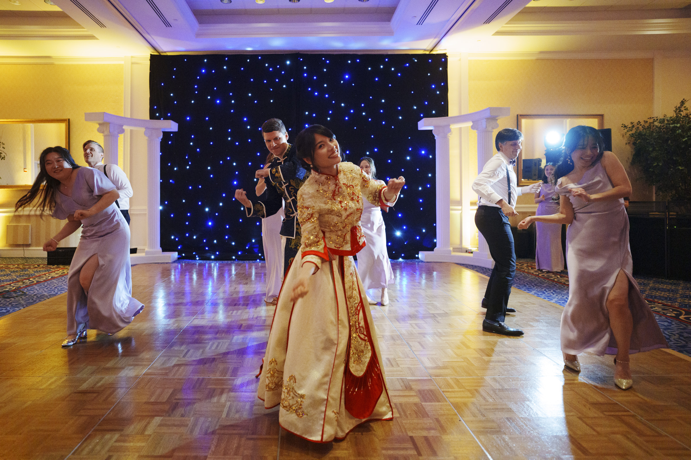 A group of people in formal attire are dancing energetically on a wooden floor. Behind them is a backdrop of twinkling lights. A woman in a traditional dress is at the center, and the others wear suits and gowns. The atmosphere is joyful and festive.