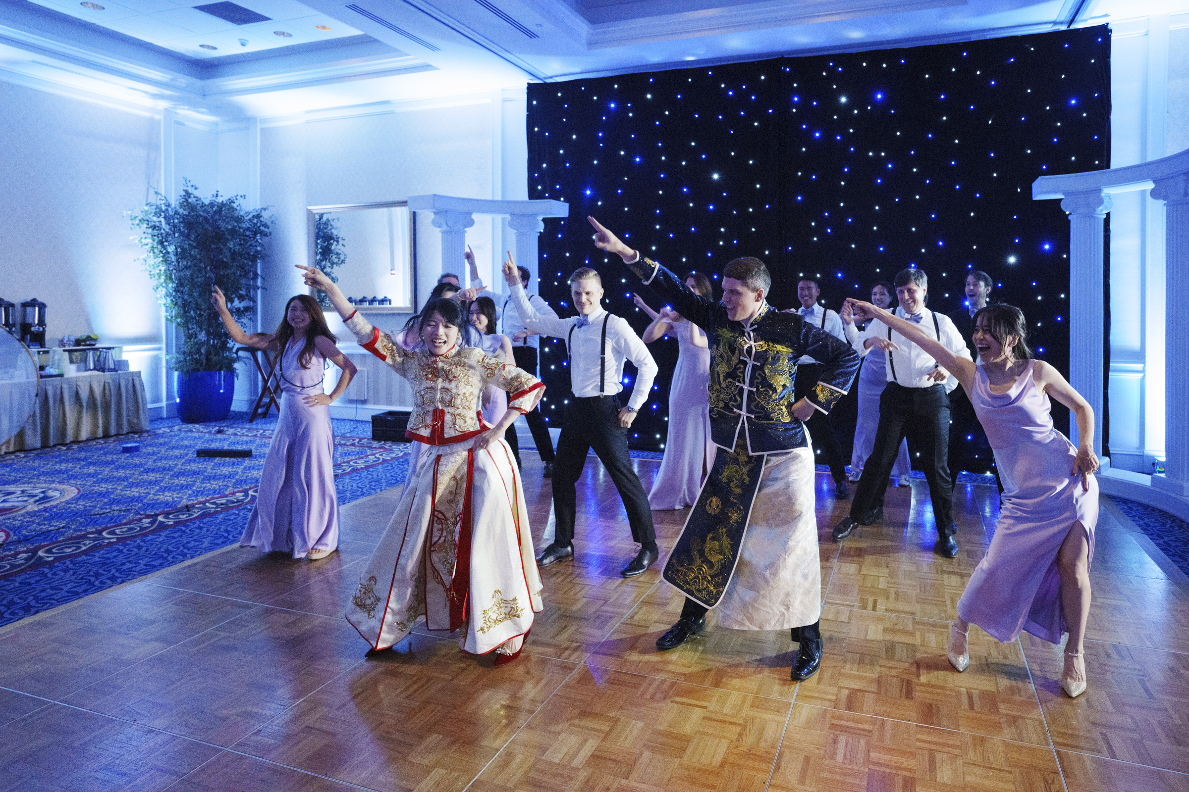 A group of people performs a choreographed dance on a wooden floor in a ballroom. They are dressed in a mix of elegant attire, including traditional and formal clothing, with a backdrop of twinkling star-like lights.