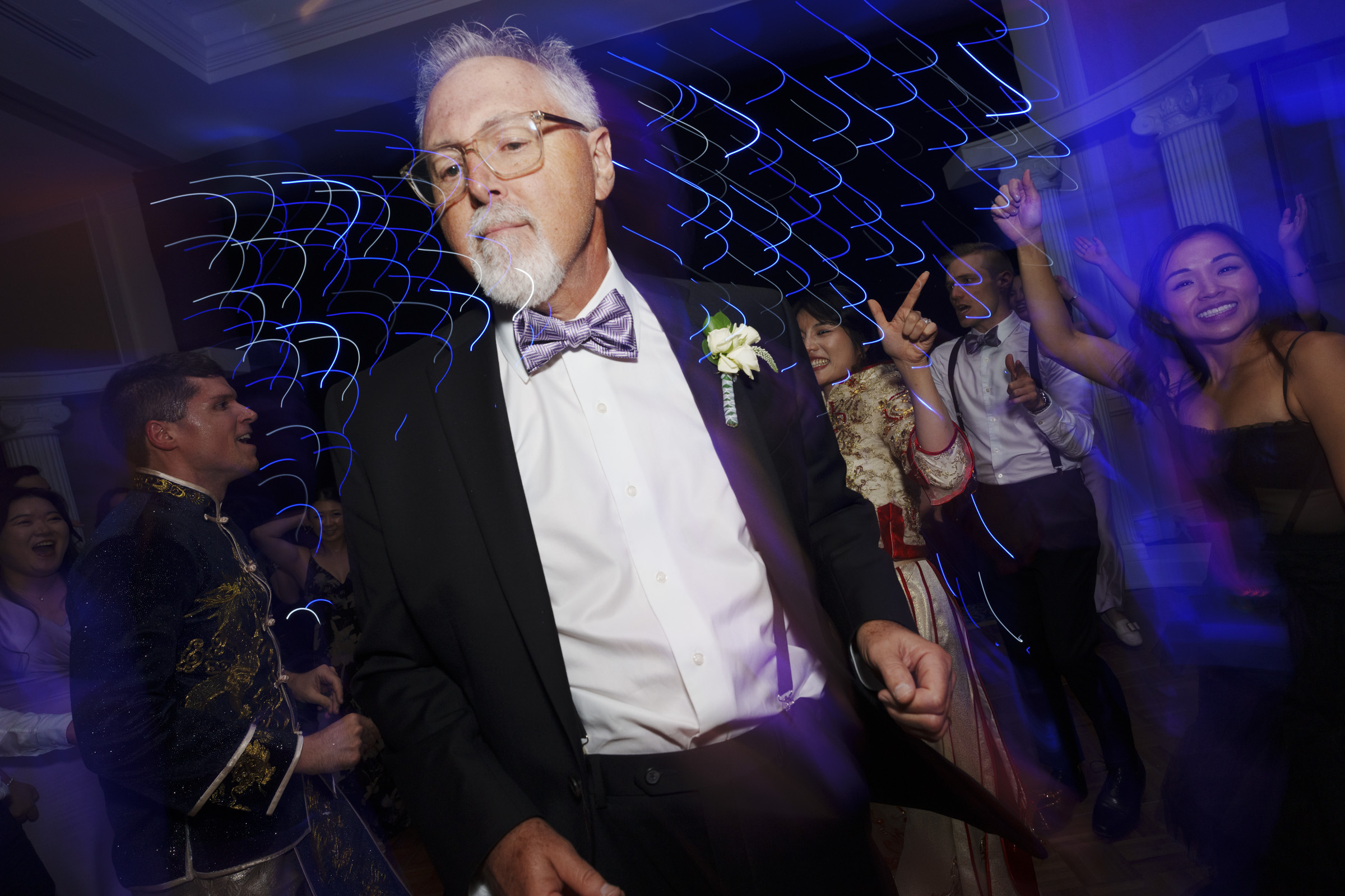 A man in a suit and bow tie dances energetically at a party. The background features vibrant, blurred lights and other people dancing and enjoying the event.