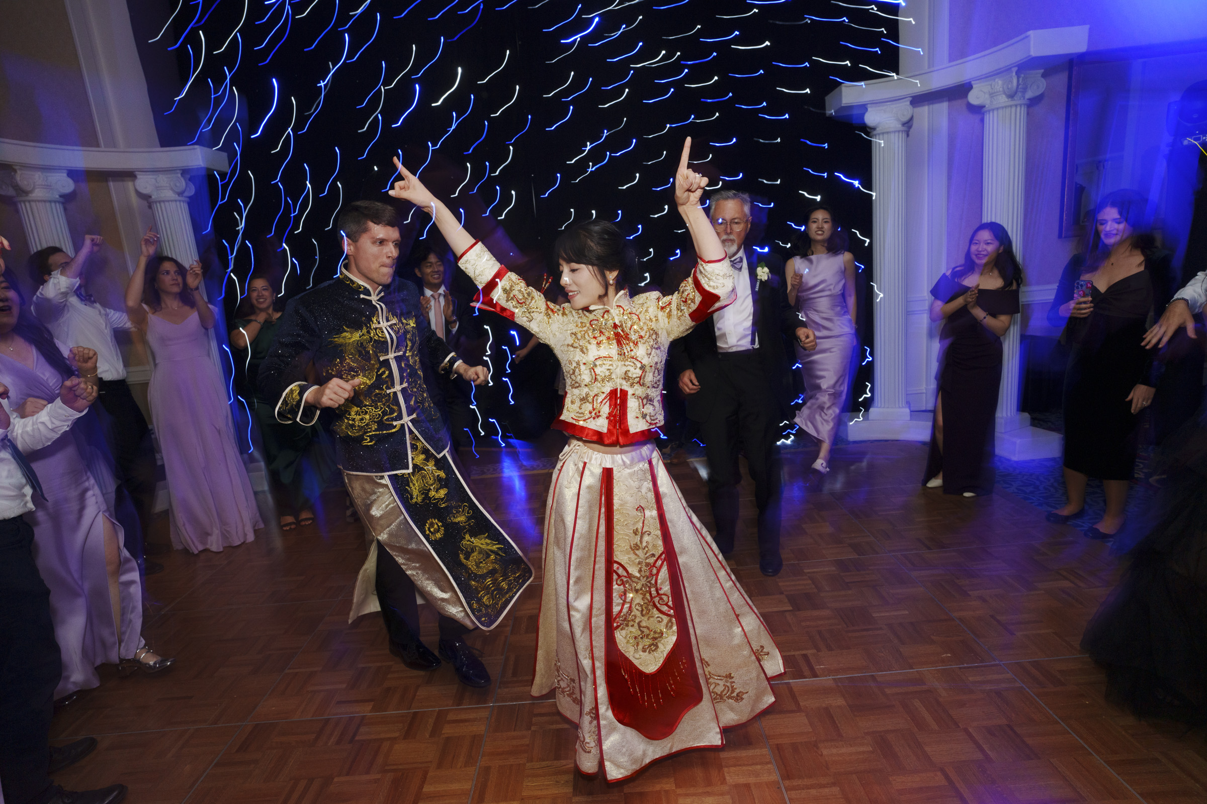 A couple is dancing energetically on a wooden floor, surrounded by guests dressed in formal attire. They wear traditional, ornate outfits, and the background features a curtain of blue lights. The atmosphere is lively and festive.