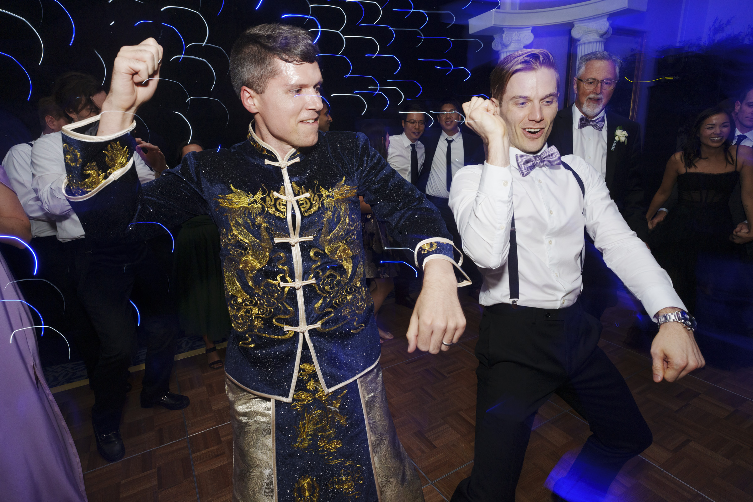 Two people dancing energetically. One is wearing a blue and gold traditional outfit, and the other is in a white shirt with suspenders and a bow tie. They are surrounded by people dressed formally, with blurry lights in the background.