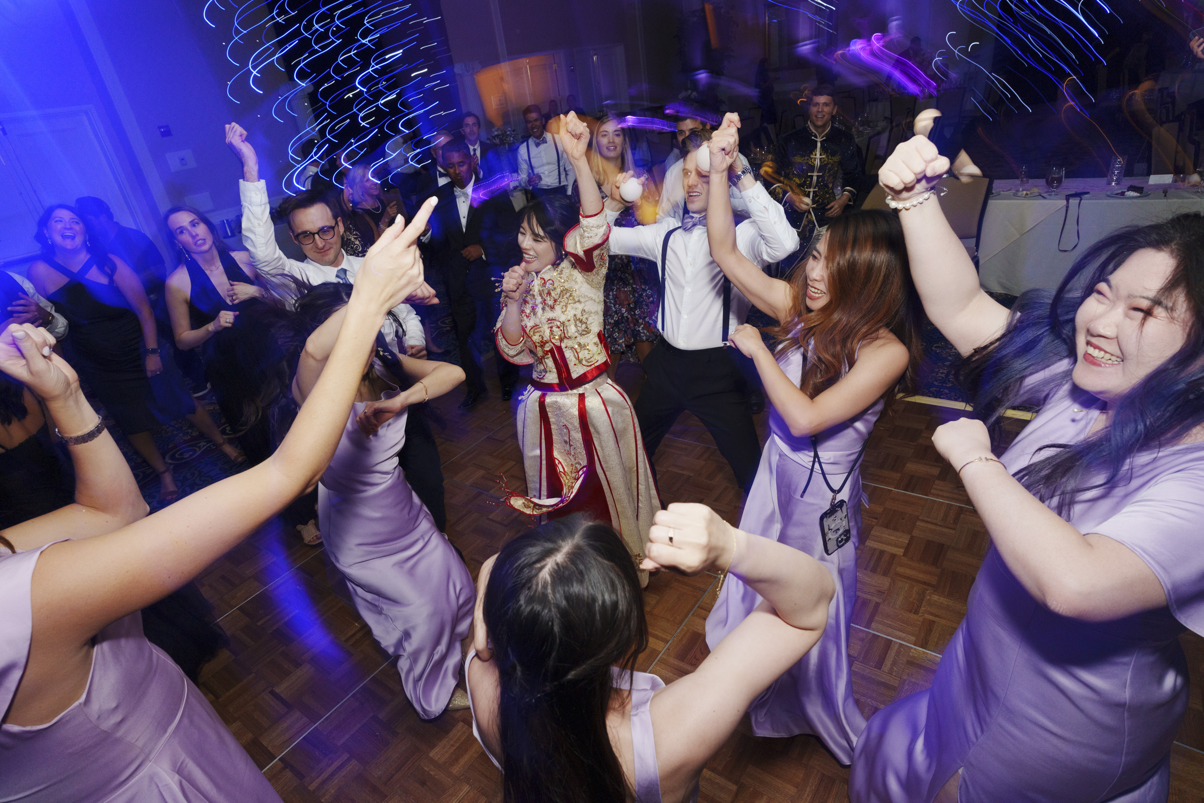 A lively dance floor scene with people in festive attire enthusiastically dancing. The room is illuminated with blue and purple lights, creating a vibrant atmosphere. A person in a distinct red and gold outfit stands at the center.