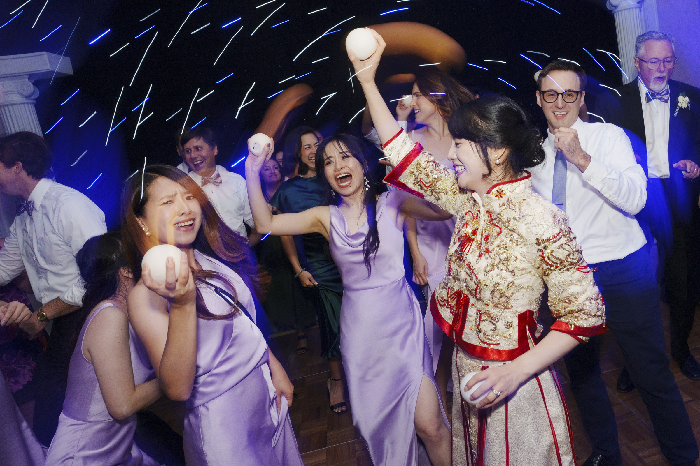 A lively party scene with people dancing. A woman in traditional attire and several others in lilac dresses hold lights, creating streaks in the air. Everyone appears joyful and engaged in celebration. Blue and purple lighting enhances the festive atmosphere.