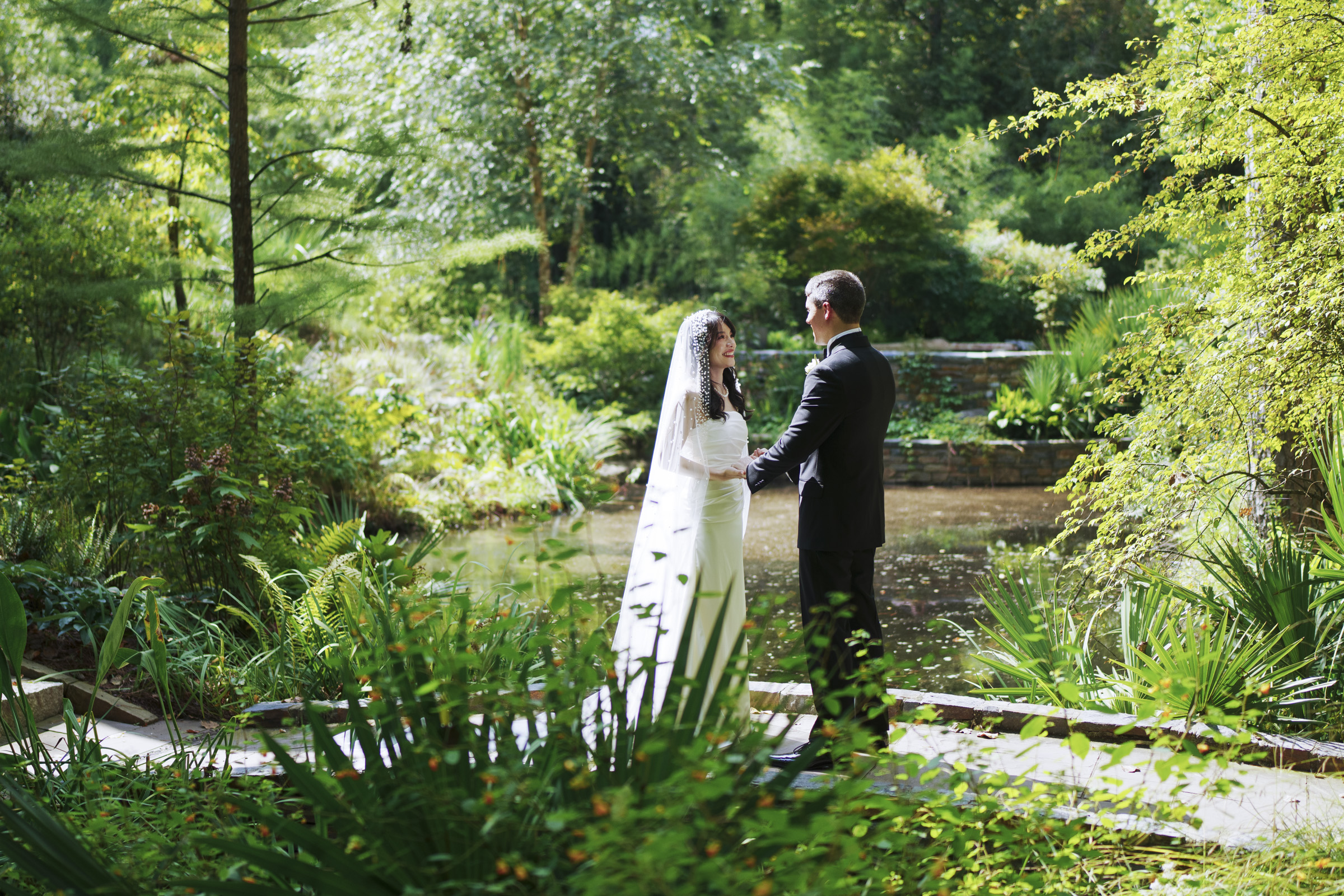 A bride and groom stand on a pathway in a lush garden, holding hands and smiling at each other. The scene is peaceful, with greenery and a small pond in the background, surrounded by sunlight filtering through the trees.