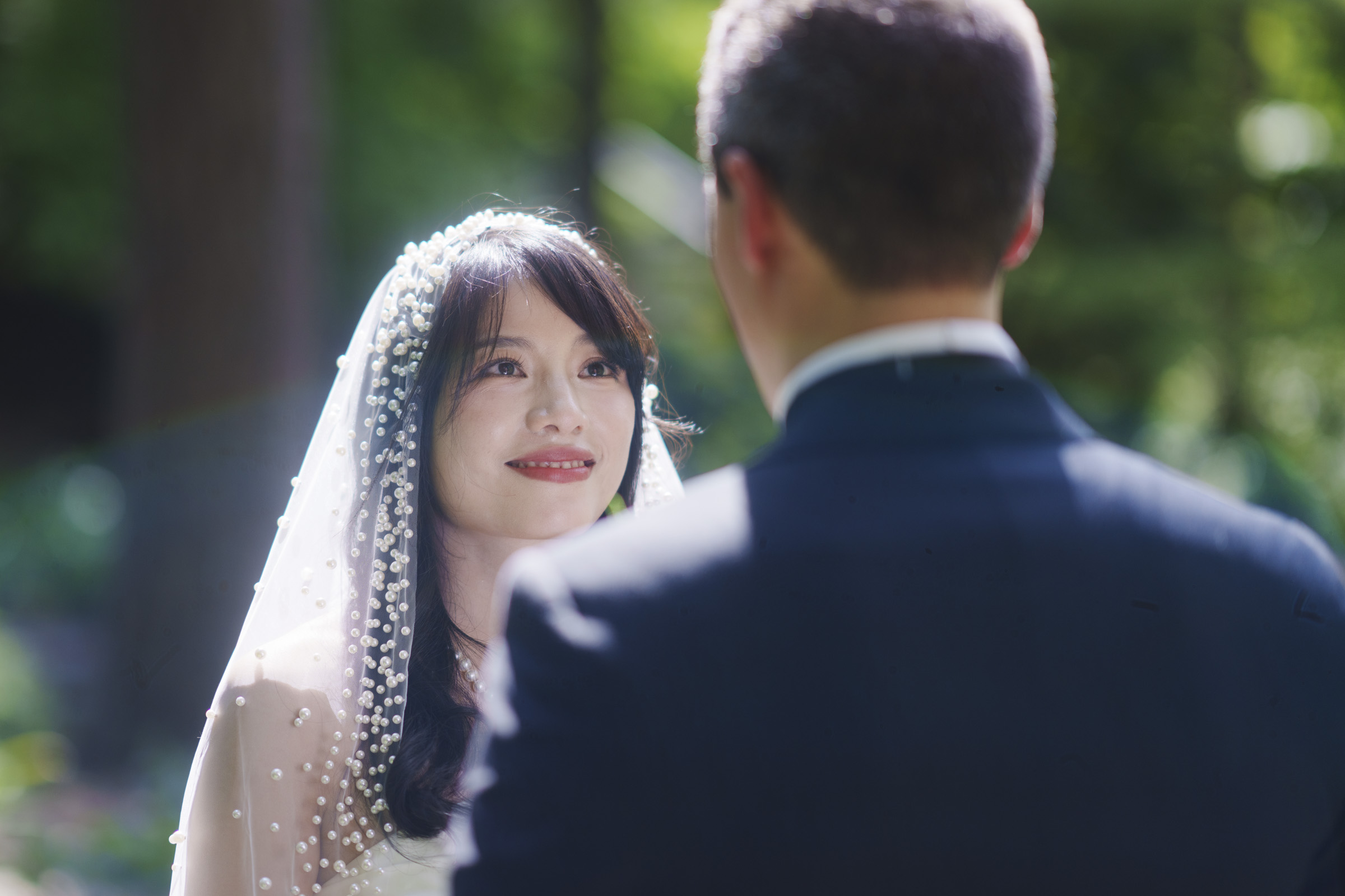 A bride and groom stand facing each other outdoors. The bride wears a pearl-studded veil and smiles, looking at the groom, who wears a dark suit. Sunlight filters through green trees in the background, creating a warm, romantic ambiance.