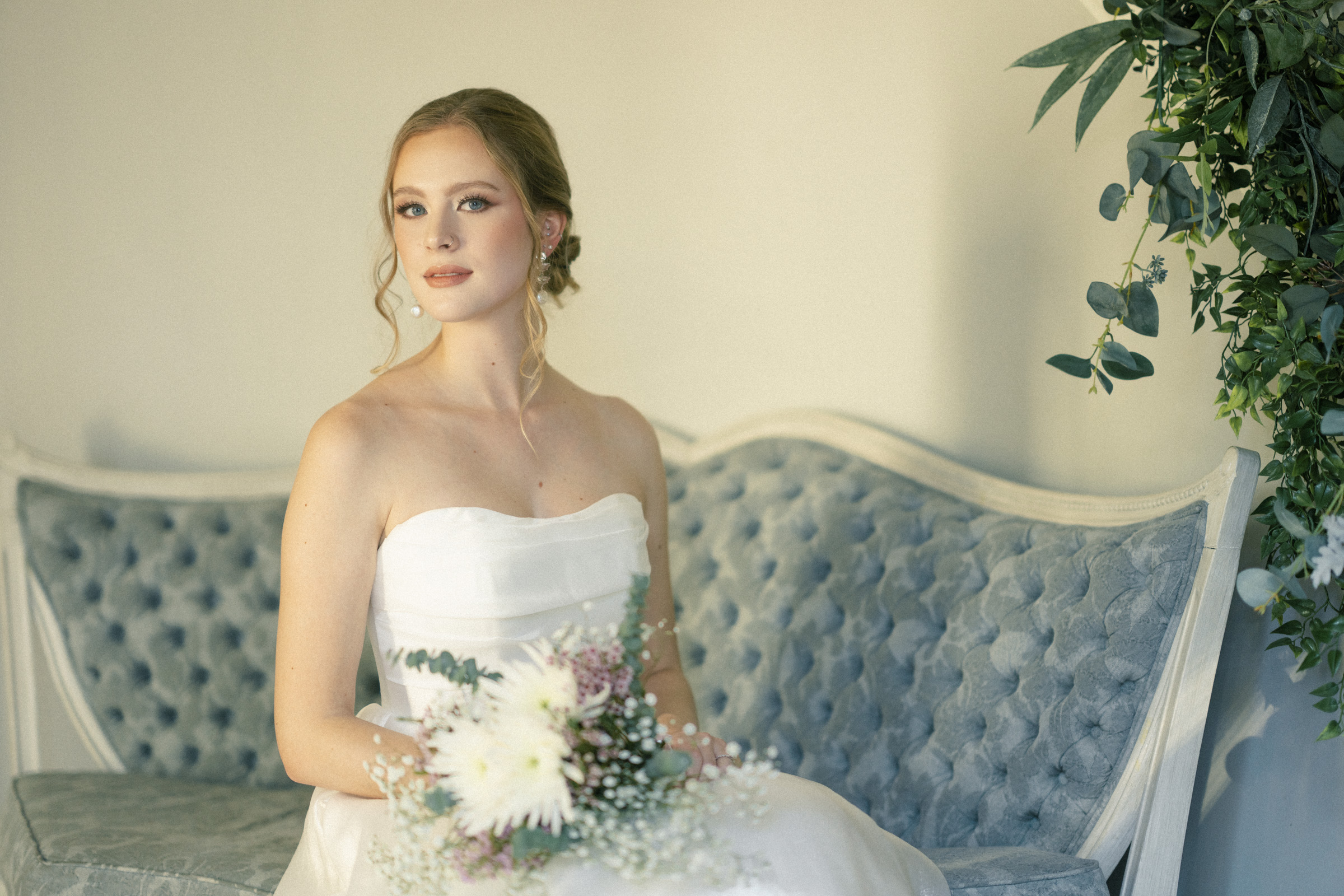 A bride in a strapless white gown sits on a light blue tufted bench, holding a bouquet of white and pink flowers. Her hair is styled in an updo, and she gazes softly. The background includes green leafy plants.