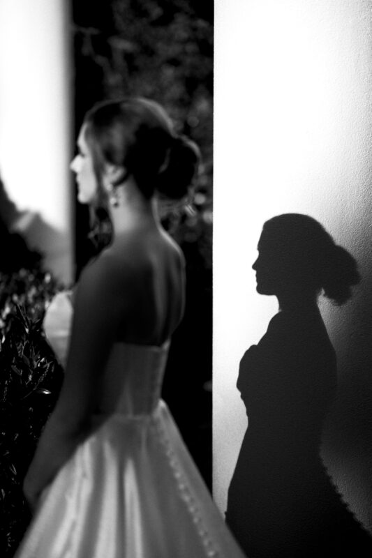 A woman in a wedding dress stands against a light-colored wall, casting a shadow. The scene is in black and white, with her shadow creating a silhouette that mirrors her profile and elegant gown.