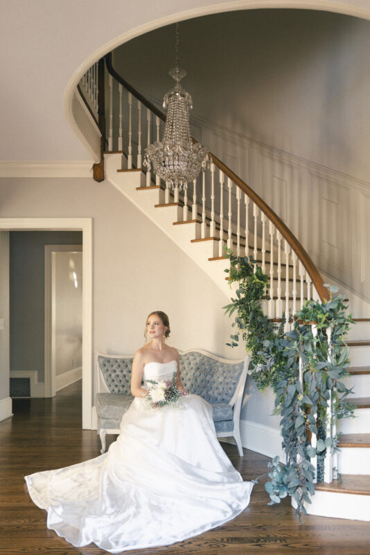 A bride in a white gown sits on a blue upholstered bench under a grand, curved staircase adorned with greenery. A chandelier hangs above, and she holds a bouquet, gazing thoughtfully to the side. The setting is elegant and serene.