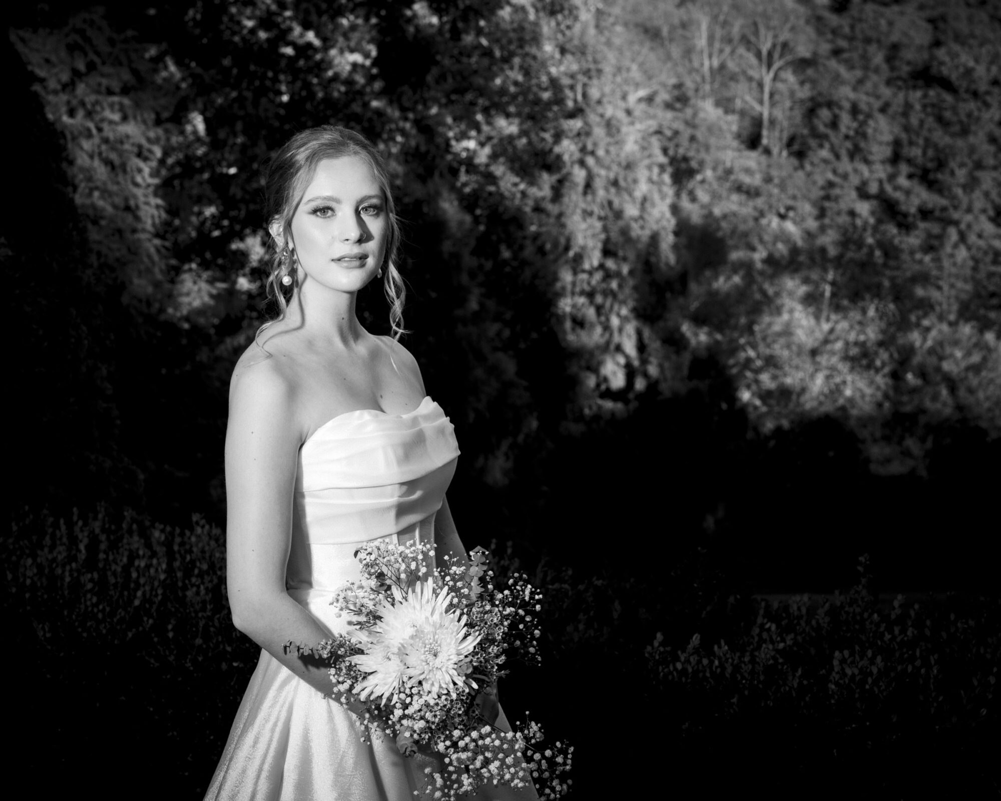 A bride in a strapless gown holds a bouquet and stands in a garden. The image is in black and white, highlighting the contrast between her dress and the lush background. She gazes into the distance, with trees softly blurred behind her.