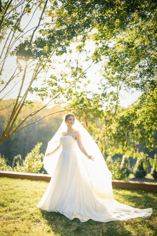 A bride in a white wedding gown stands outdoors, smiling under the shade of leafy trees. Sunlight filters through the branches, highlighting her veil as it flows in the gentle breeze. The scene is set against a backdrop of greenery and soft sunlight.