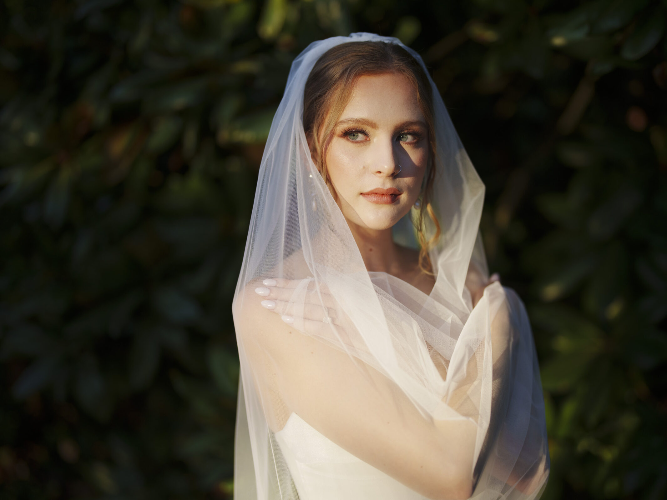 A bride stands in front of a leafy backdrop, wrapped in a translucent veil. She wears a white strapless gown and gazes softly into the distance, with soft sunlight casting a warm glow on her face.