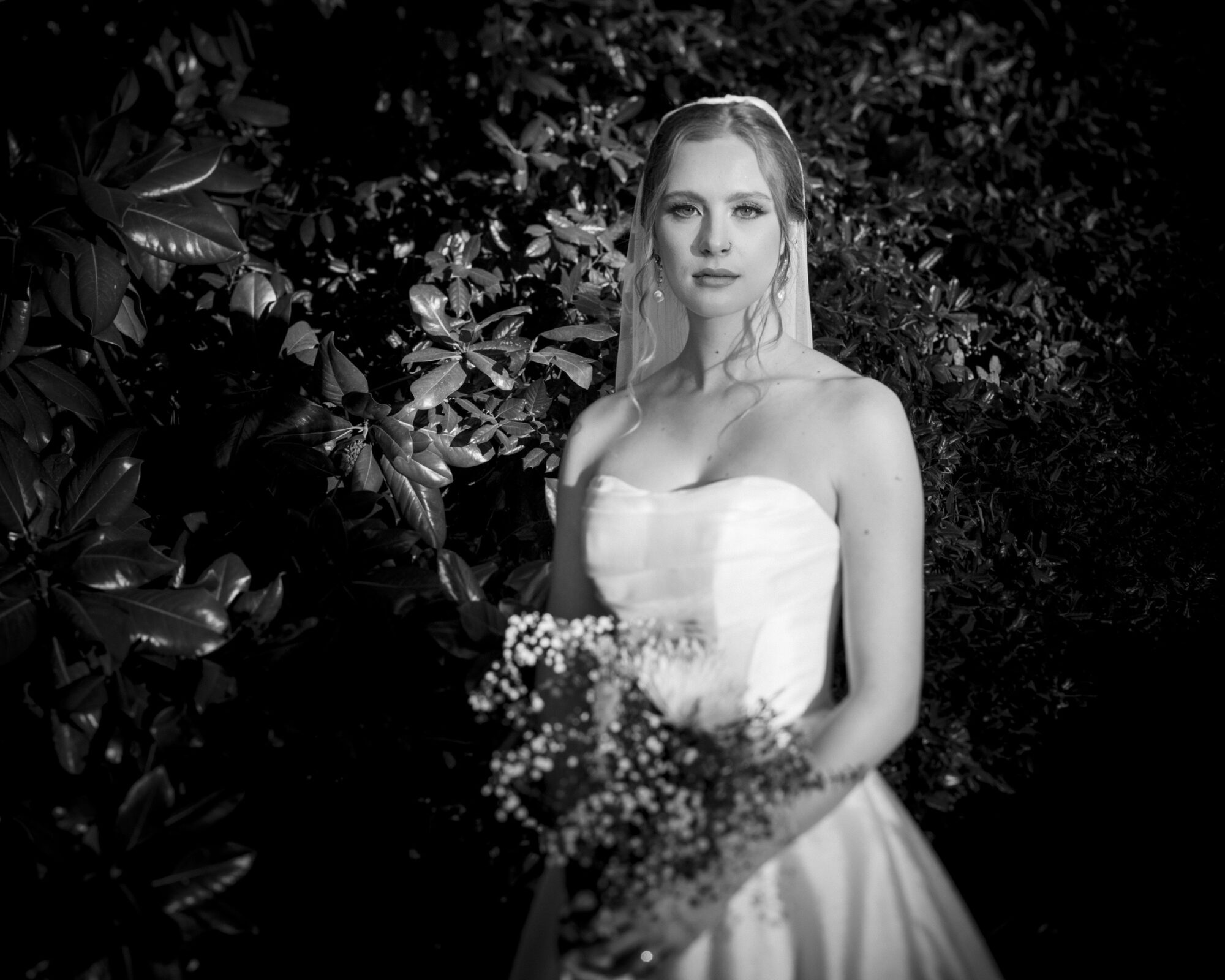 A bride in a strapless wedding dress stands in front of lush foliage. She holds a bouquet of flowers and wears a veil. The image is in black and white, highlighting her serene expression and the gowns elegant details.