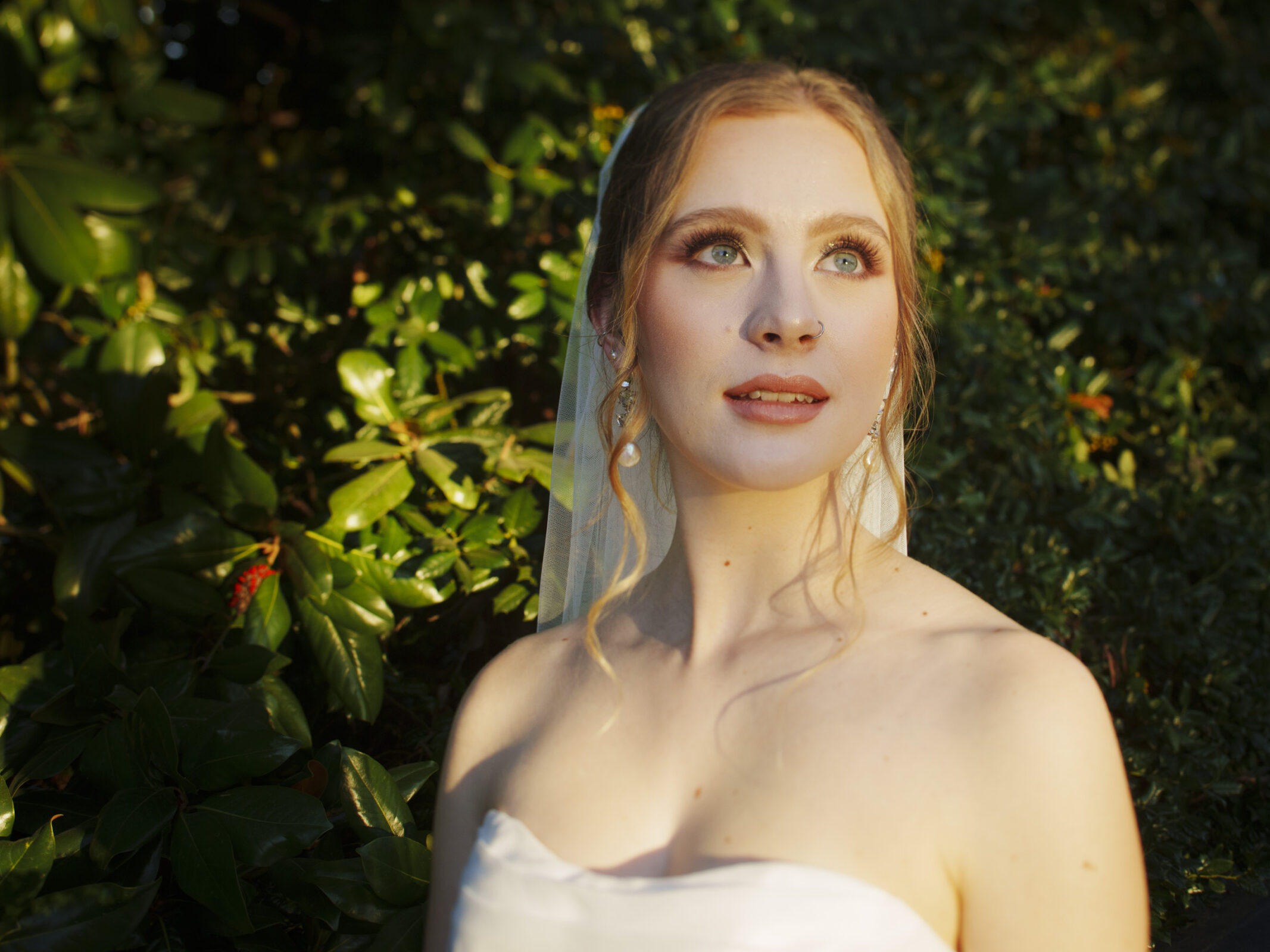 A woman wearing wedding attire stands outdoors, bathed in warm sunlight. She is in front of lush green foliage, looking slightly upward with a contemplative expression. Her hair is elegantly styled, and she wears a veil.