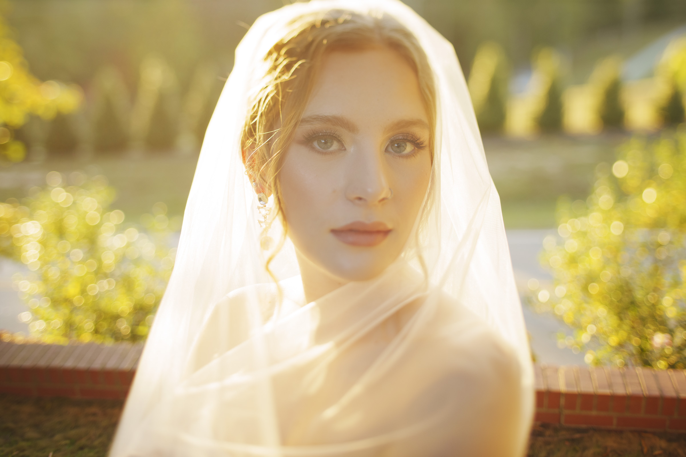 A bride in a white veil stands outdoors in soft, golden sunlight. She has light makeup and her hair is styled in loose curls. The background is blurred, showcasing greenery and a brick wall.