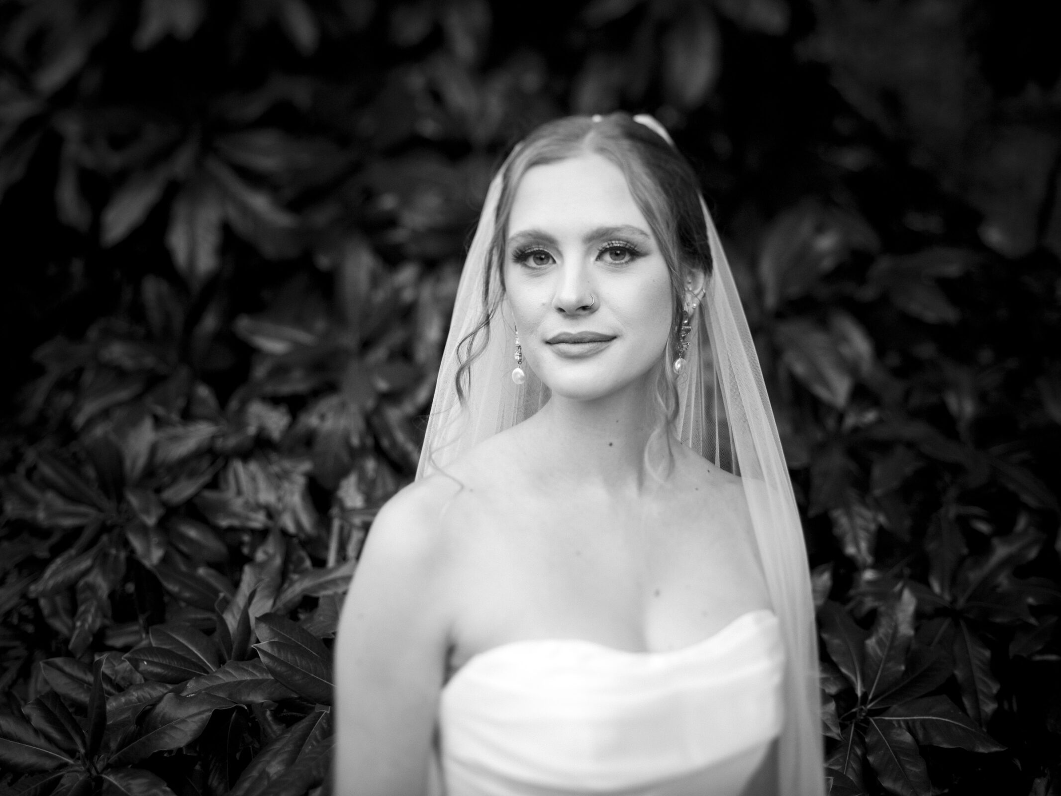 A bride in a strapless wedding dress with a long veil stands in front of a foliage background, looking at the camera. The image is in black and white, highlighting her subtle smile and elegant earrings.