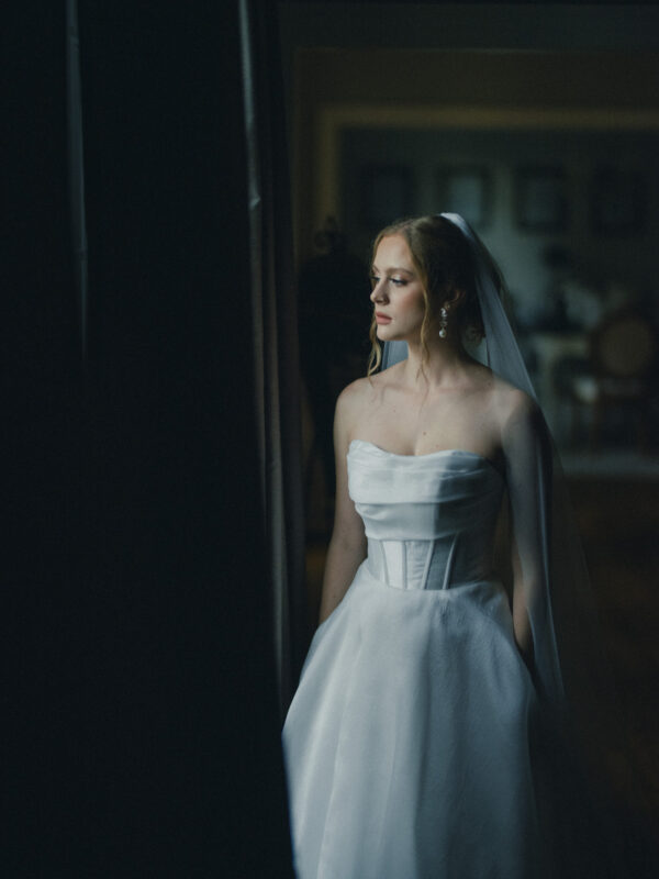 A bride in a white strapless gown and veil stands by a window, gazing outside. The room is softly lit, creating a tranquil and reflective atmosphere.