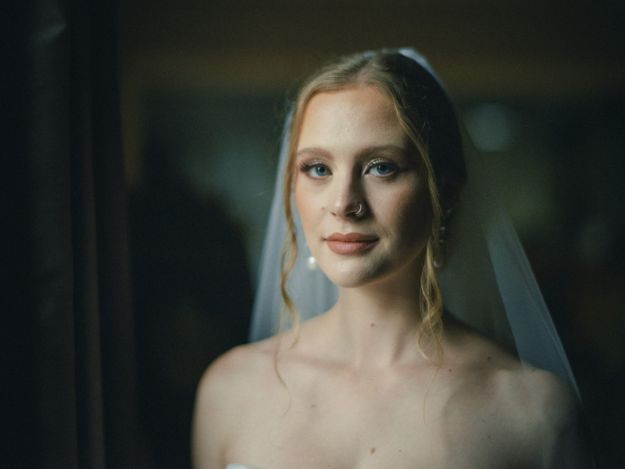 A bride with long, blonde hair and a sheer veil looks towards the camera. She is wearing subtle makeup and a strapless gown, with soft lighting emphasizing her serene expression.