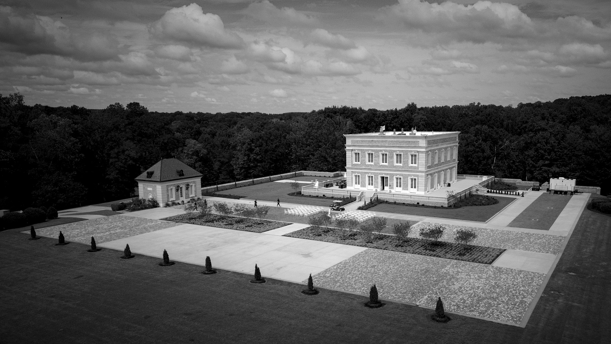 Aerial view of the elegant Oxbow Estate, ideal for wedding photography, with a large mansion surrounded by manicured gardens and a spacious paved courtyard. The main building is two stories with a flat roof, flanked by a smaller annex. Dense trees form the background under a cloudy sky.