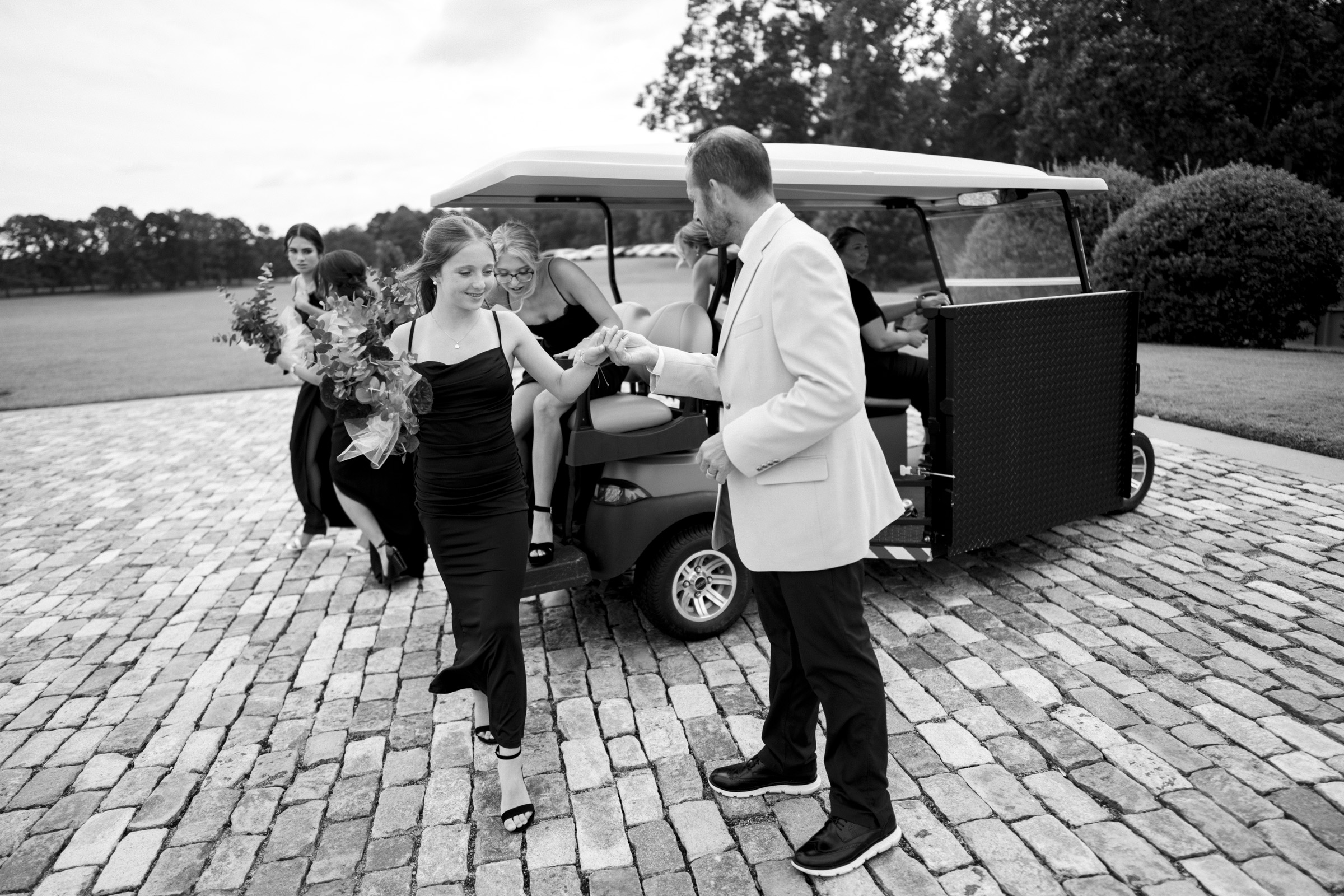 A black-and-white image captures a person in a suit assisting another in a dress as they step from a golf cart, emblematic of Oxbow Estate Wedding Photography. Nearby, guests mingle on the cobblestone path amid verdant fields and towering trees.