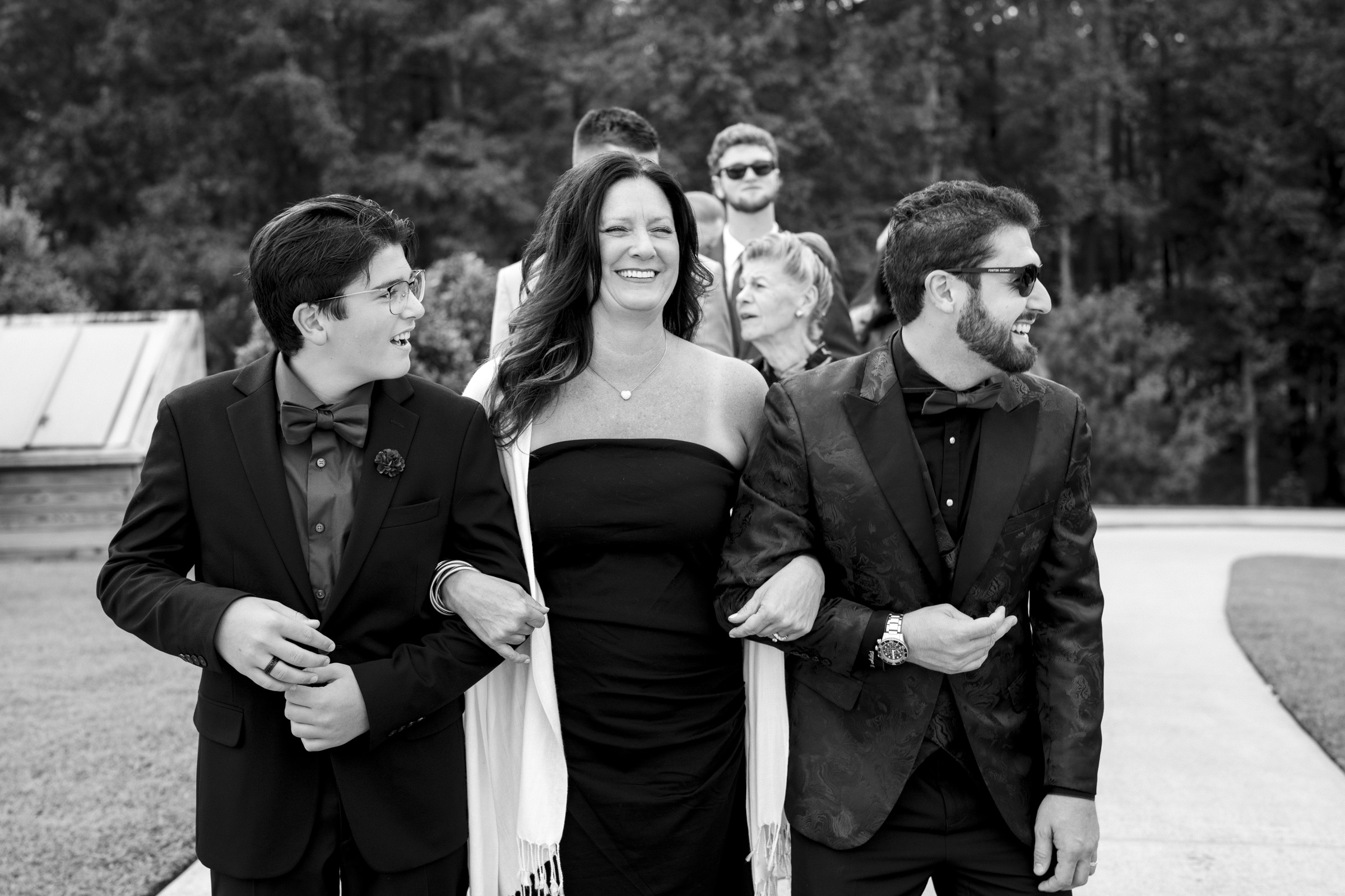 A black-and-white image captures the joy of a group strolling outdoors, smiling and chatting. In the foreground, a woman in a dress is arm-in-arm with two men in suits and bow ties. Other guests from the Oxbow Estate wedding are visible amidst the trees lining the pathway.