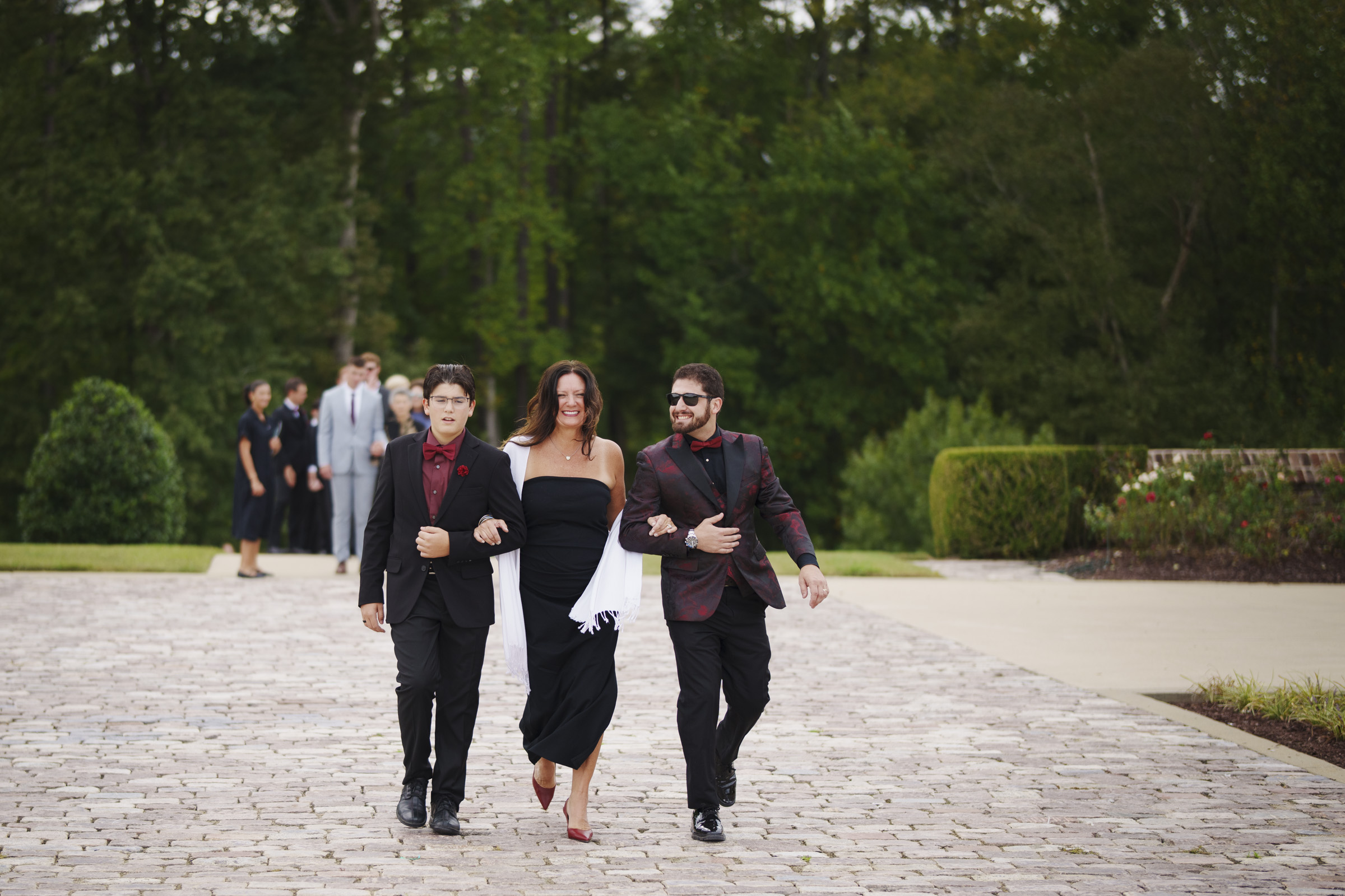 At the picturesque Oxbow Estate, three people in formal attire stroll along a stone walkway. The woman in a black dress is flanked by two men in matching dark suits with red accents. Lush trees frame the scene, while a distant group of guests adds to the elegant wedding atmosphere.