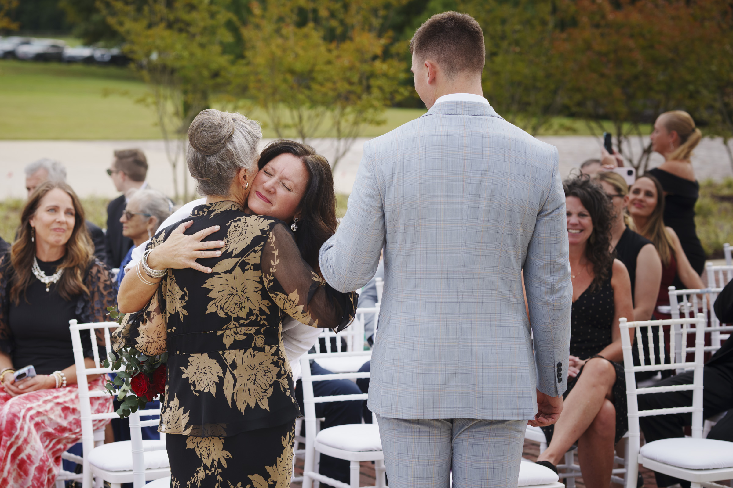 At an enchanting Oxbow Estate wedding, a woman in a floral dress embraces another woman at an outdoor event, while a man in a light gray suit stands beside them. Guests seated on white chairs smile as they watch this joyful interaction, surrounded by lush greenery and a winding pathway.