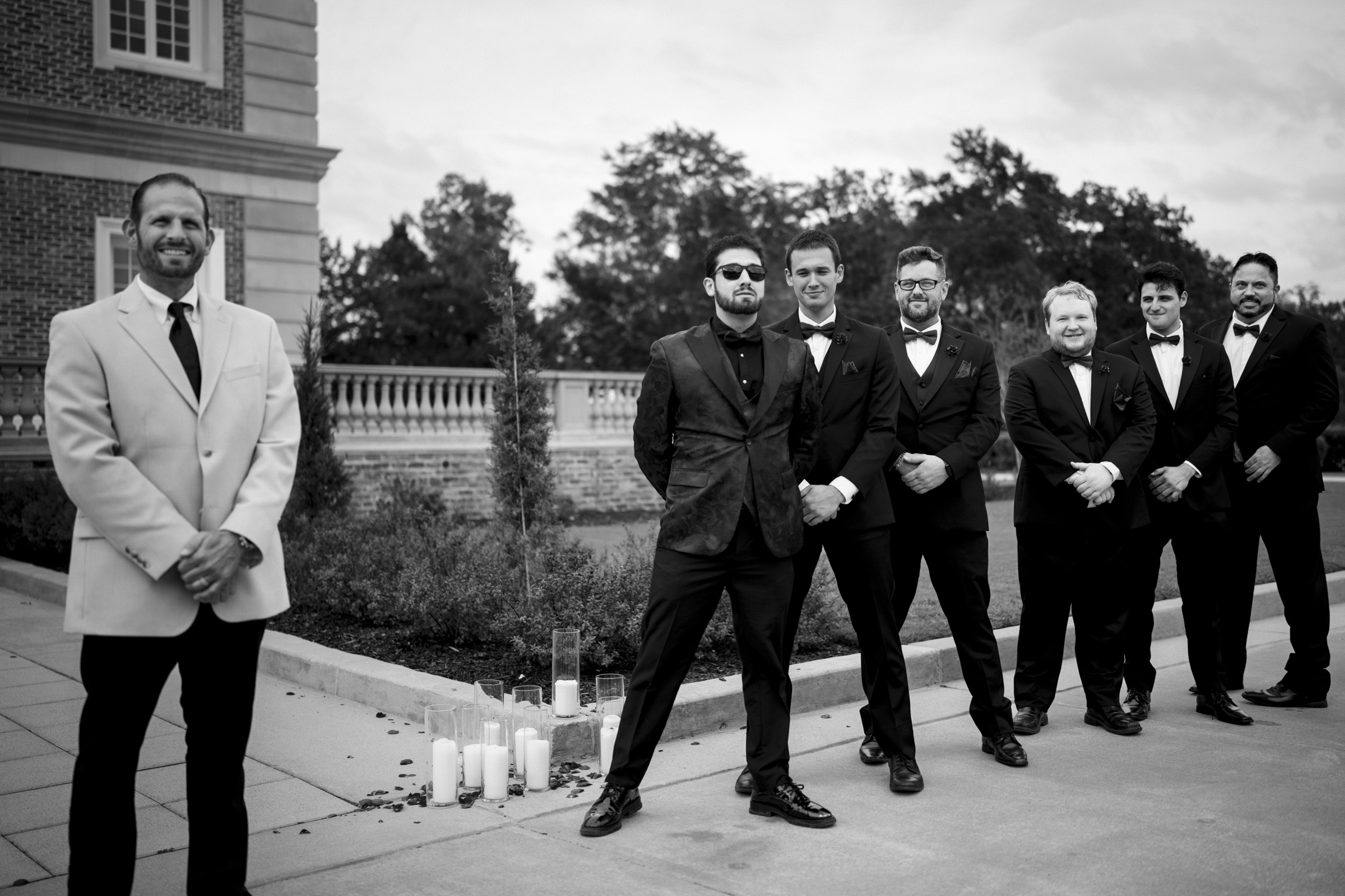 A group of six men in formal attire stand outdoors at Oxbow Estate, with a lined row of candles on the ground. One man, wearing a light-colored jacket, stands separated on the left. The overcast sky and visible trees frame this elegant wedding photography moment.