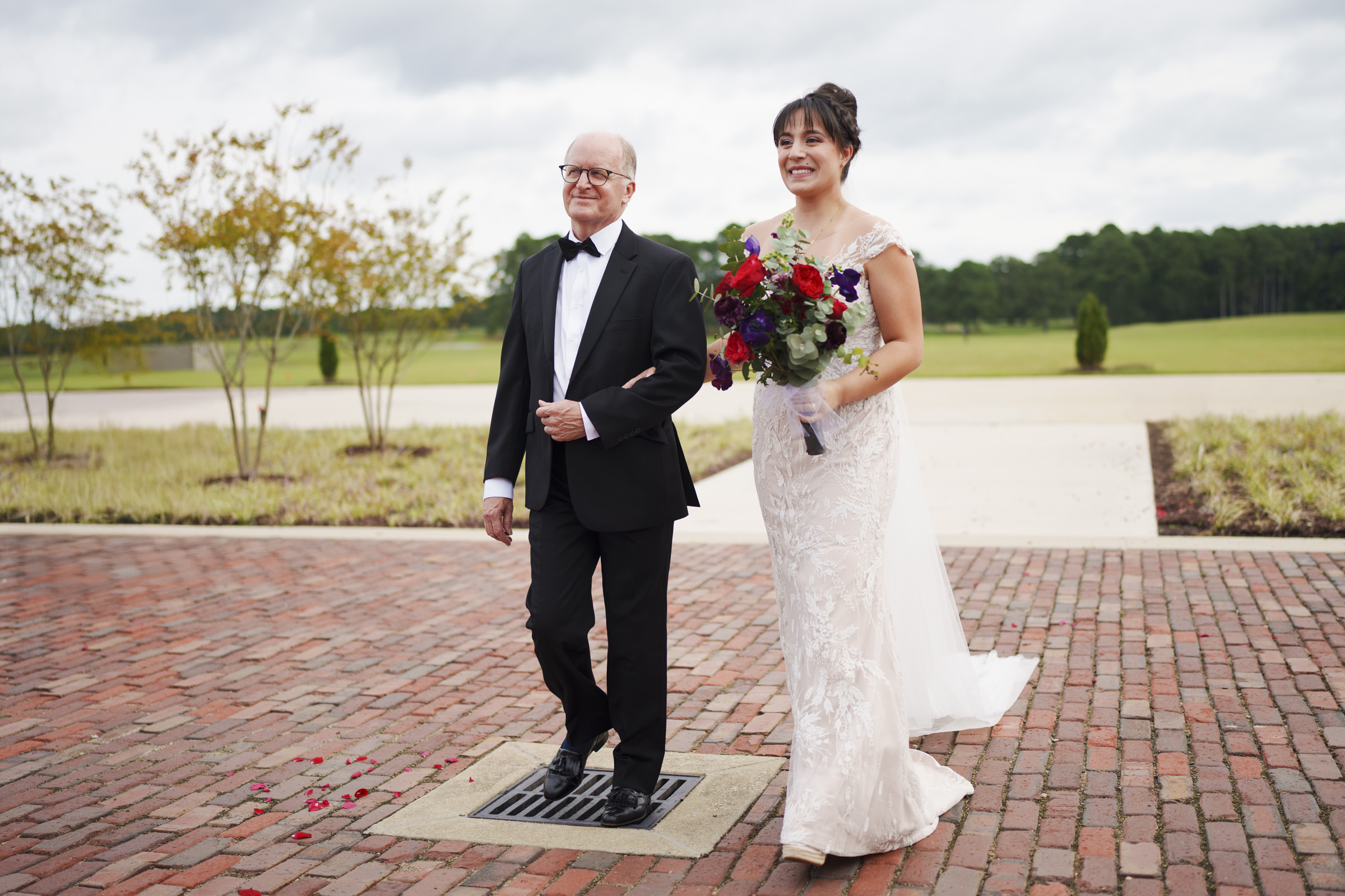 A bride in a white lace gown holds a bouquet of red and white flowers while walking arm-in-arm with an older man in a black suit and bow tie on a brick path at Oxbow Estate, with the grassy landscape captured beautifully in this wedding photography.