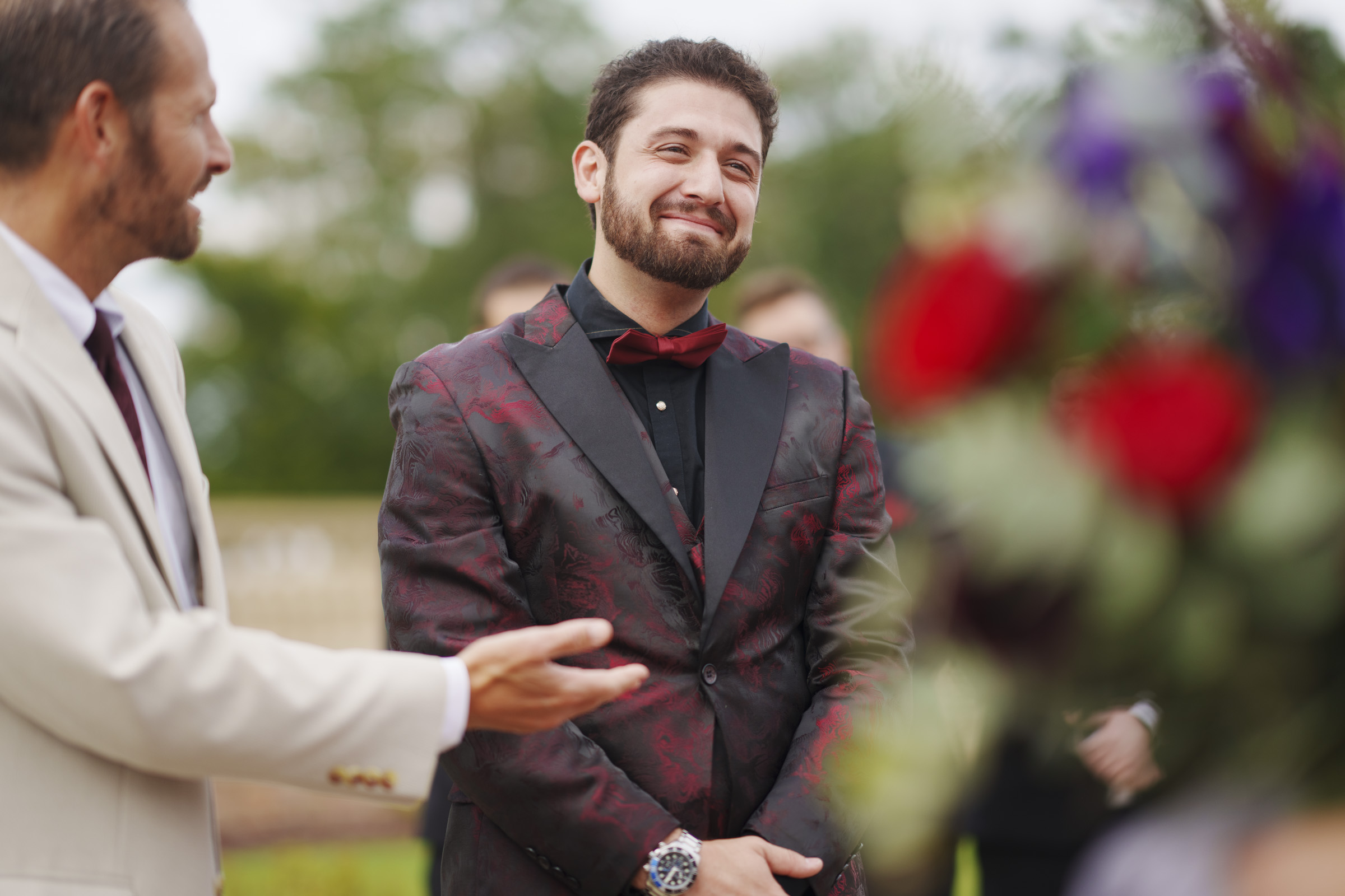 At Oxbow Estate, a man in a dark patterned suit and red bow tie stands smiling with his hands clasped during the wedding photography session. Another man in a light suit gestures toward him, as out-of-focus flowers and trees artfully fill the scene.