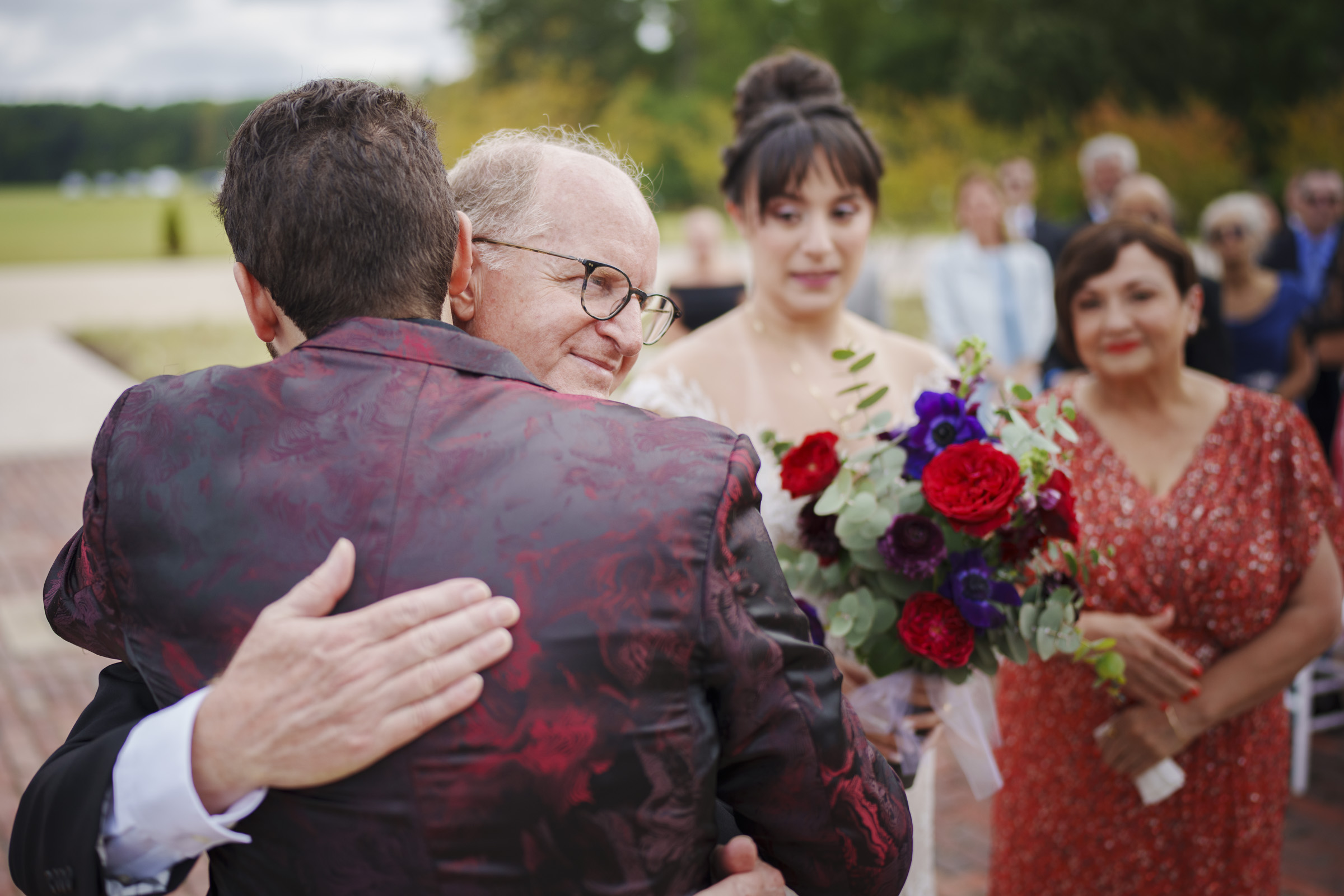 A man in a patterned suit hugs an older man at Oxbow Estate. Nearby, a woman in a wedding dress holds a colorful bouquet, while another in a red dress stands in the background. They are outdoors on a sunny day, surrounded by greenery and trees—captured beautifully through wedding photography.