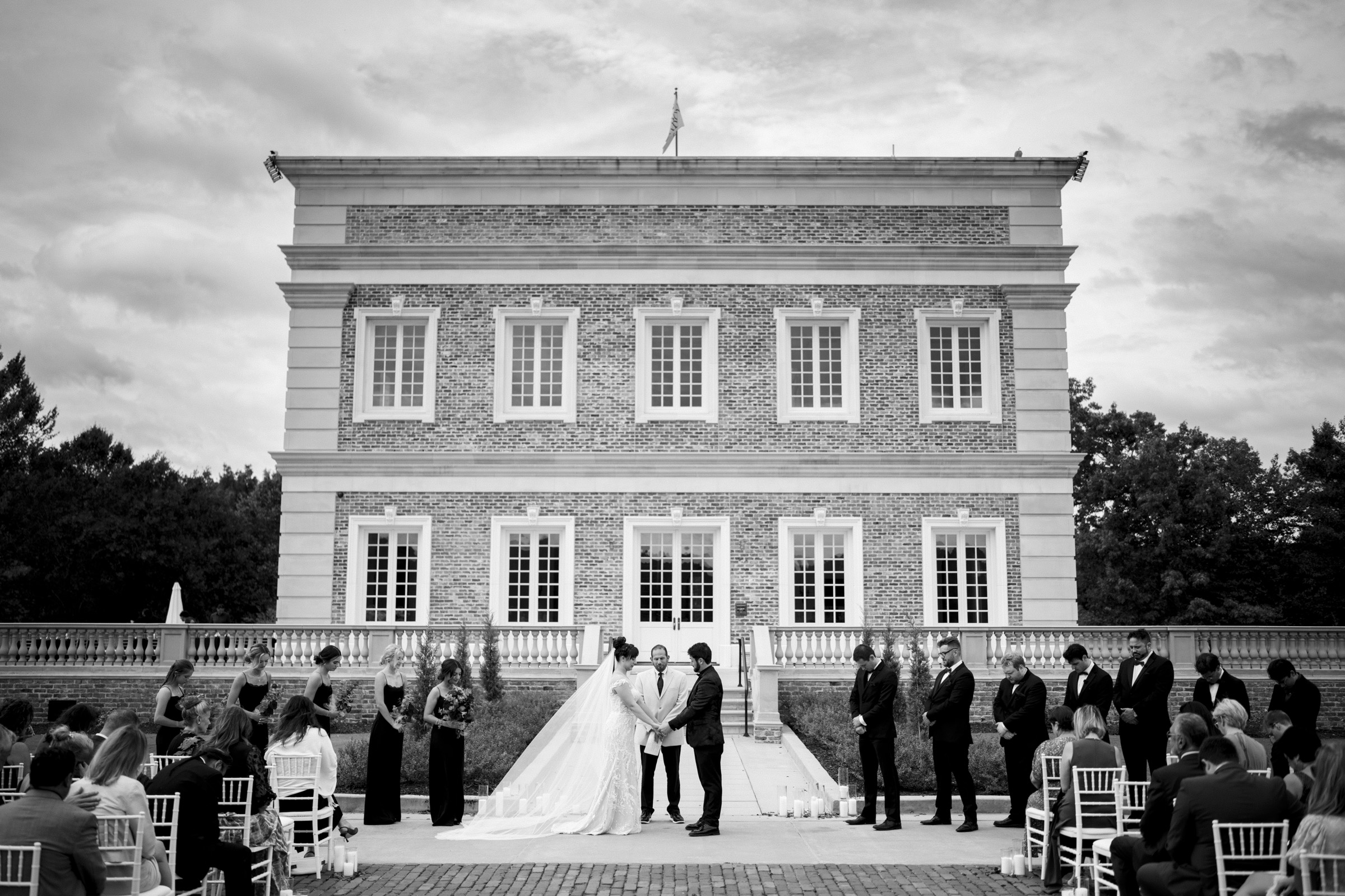 A black and white photo captures a wedding ceremony at the elegant Oxbow Estate. The bride and groom stand at the center in front of a classic brick building, surrounded by their wedding party and guests seated on either side.